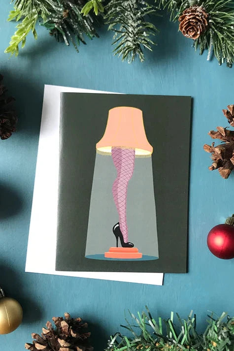 A card from a box set by Carabara Designs, featuring the iconic Leg Lamp from the movie "A Christmas Story," is shown against a teal background with holiday decor around it.