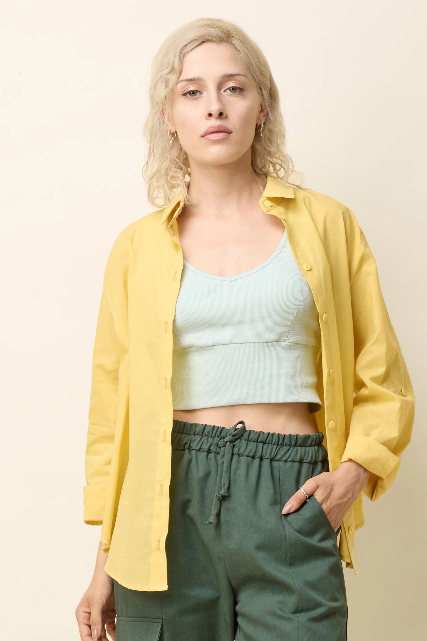 A woman wearing the Chila Shirt by Cokluch in Banana, a button down shirt with a blouse collar, rounded hem that falls below the hip, and long sleeves that can be cinched with a button and strap closure. She is wearing it with a lilac coloured crop-top and green pants and standing in front of a white background.