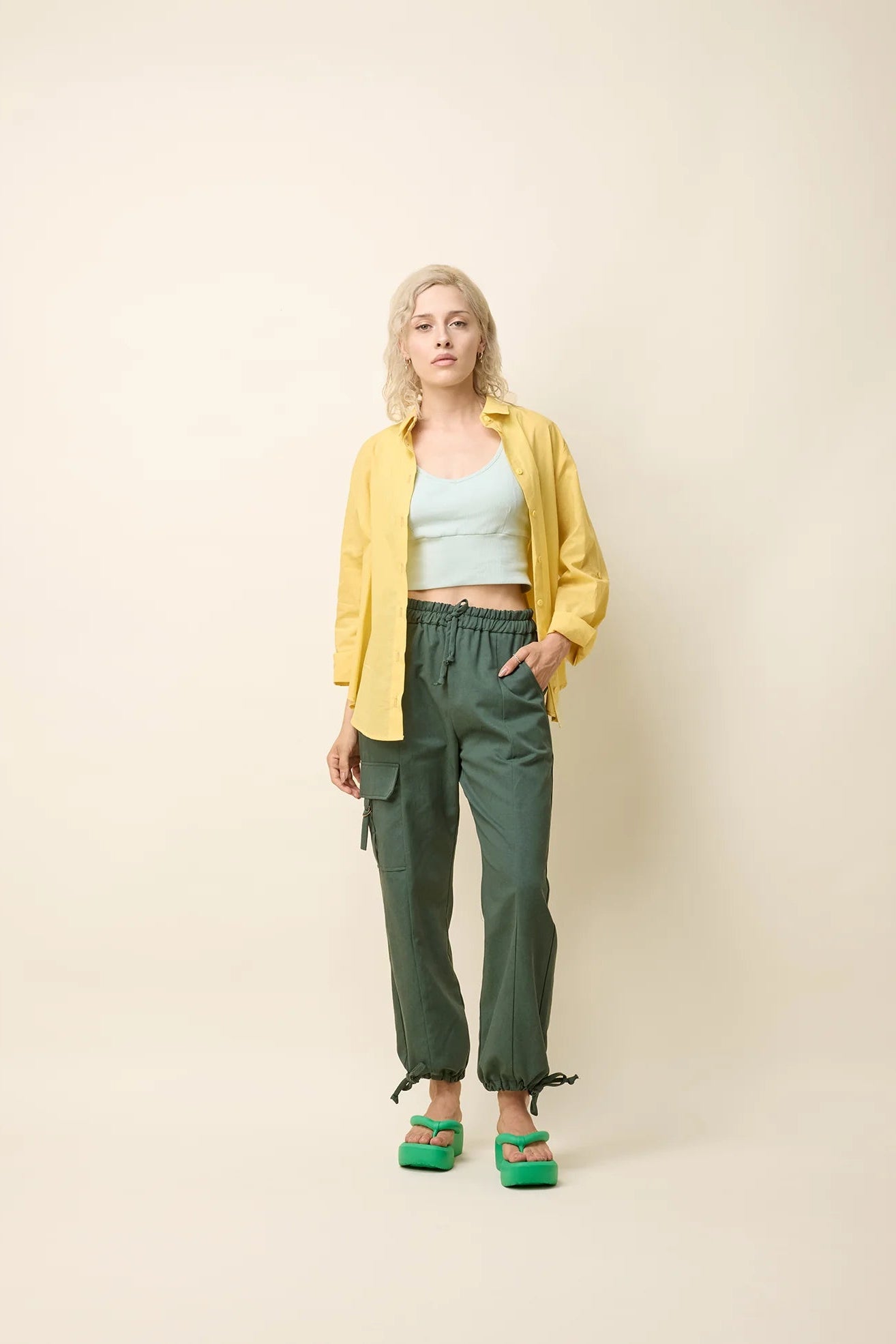 A woman wearing the Chila Shirt by Cokluch in Banana, a button down shirt with a blouse collar, rounded hem that falls below the hip, and long sleeves that can be cinched with a button and strap closure. She is wearing it with a lilac coloured crop-top and green pants and standing in front of a white background.
