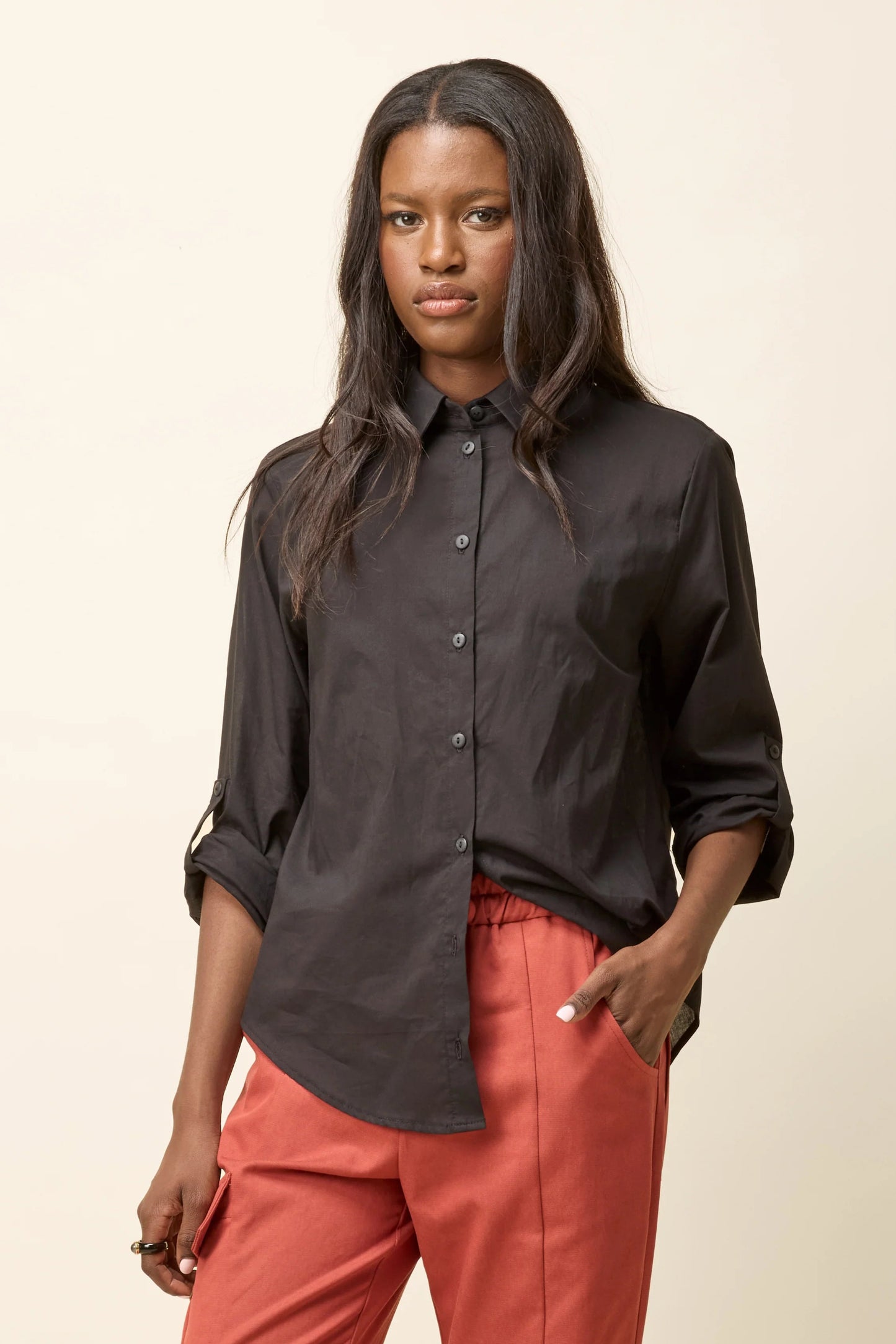 A woman wearing the Chila Shirt by Cokluch in Black, a button down shirt with a blouse collar, rounded hem that falls below the hip, and long sleeves that can be cinched with a button and strap closure. She is wearing it with orange pants and standing in front of a white background.