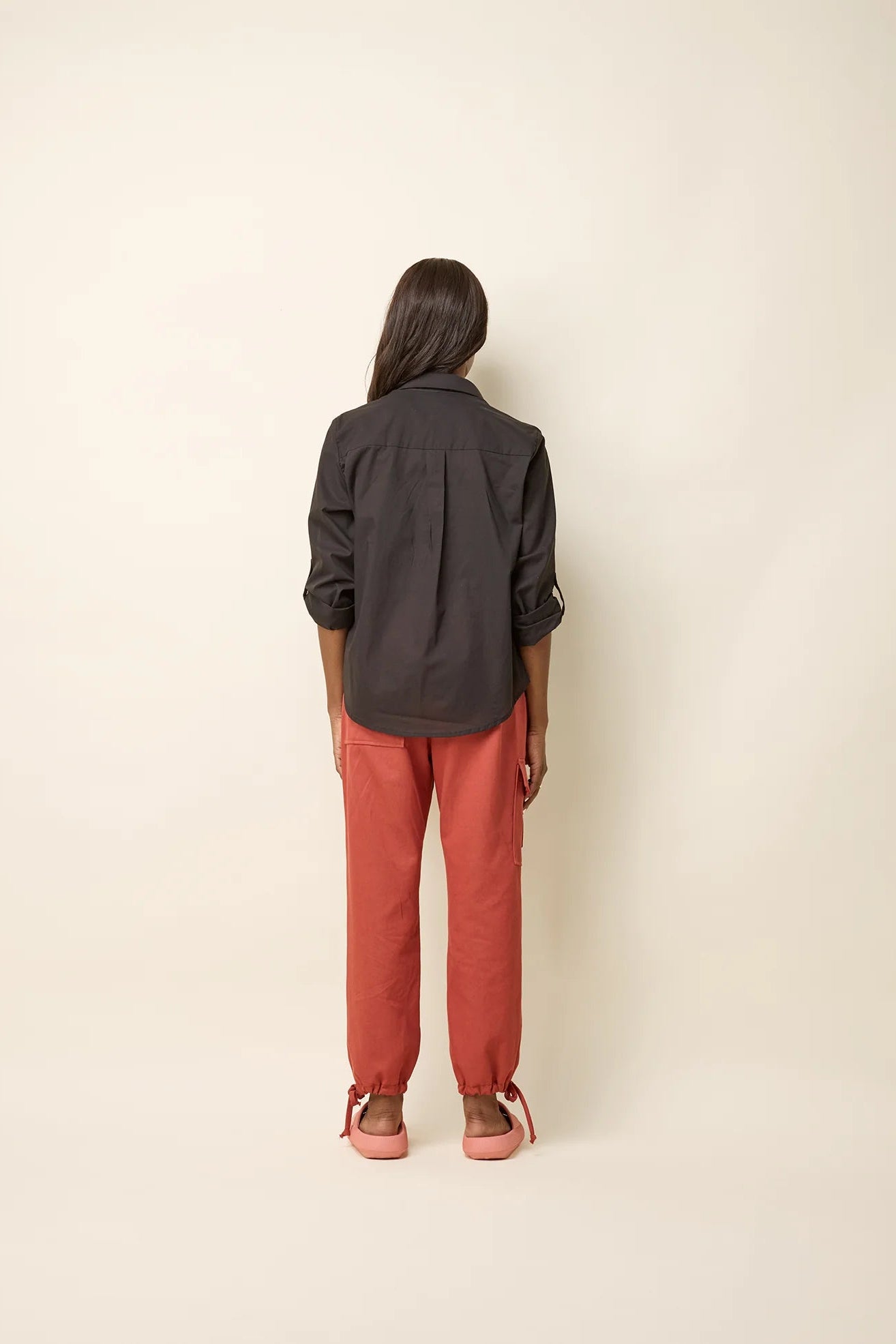 Back view of a woman wearing the Chila Shirt by Cokluch in Black, a button down shirt with a blouse collar, rounded hem that falls below the hip, and long sleeves that can be cinched with a button and strap closure. She is wearing it with orange pants and standing in front of a white background.