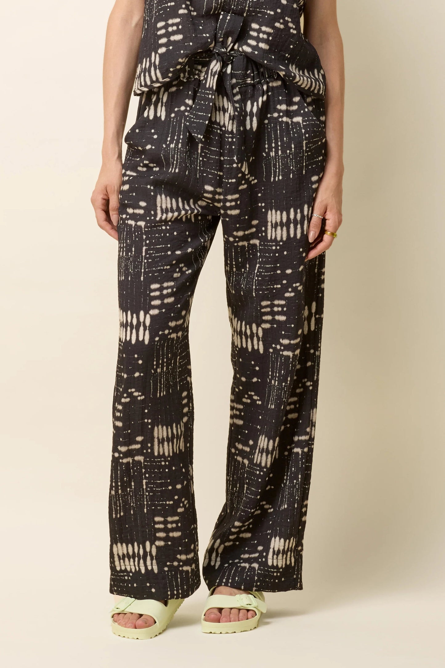 Waist down view of a woman wearing the Yael Pants by Cokluch in Black Ink print, slightly flared pants with an elastic waist, tie belt with pointed belt loops, and side pockets. She is wearing them with the matching Bayang Top and standing in front of a beige background.