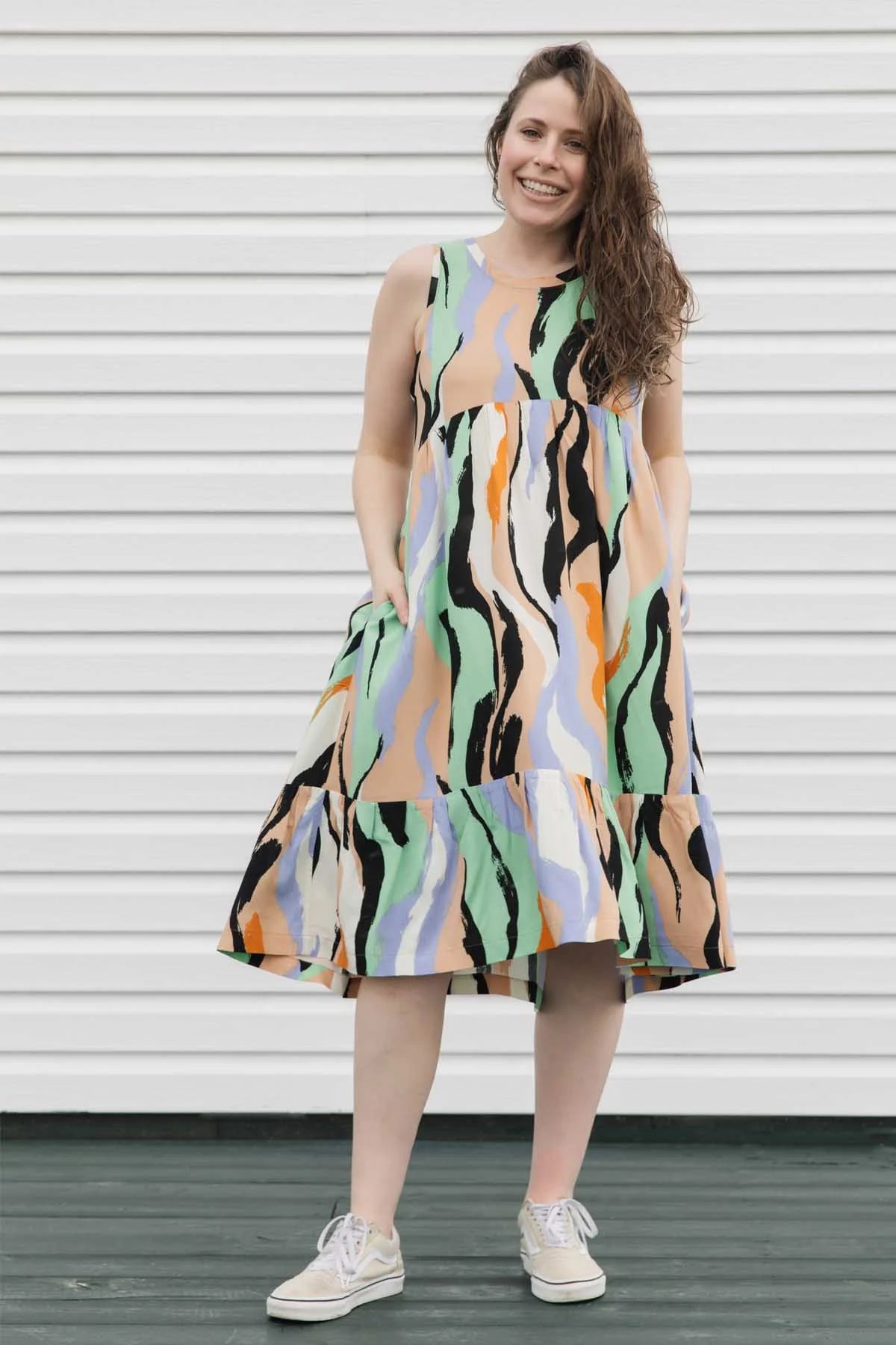 A woman wearing the Rose Dress by Copious in Mint/Peach abstract print, a sleeveless dress with an empire waist, tiered mid-calf length skirt, and pockets. She is standing against a white wall.