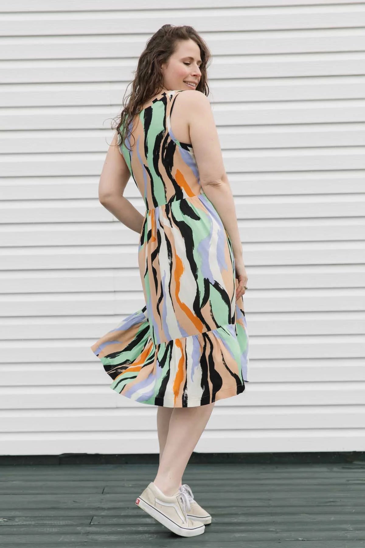 Side view of a woman wearing the Rose Dress by Copious in Mint/Peach abstract print, a sleeveless dress with an empire waist, tiered mid-calf length skirt, and pockets. She is standing against a white wall.