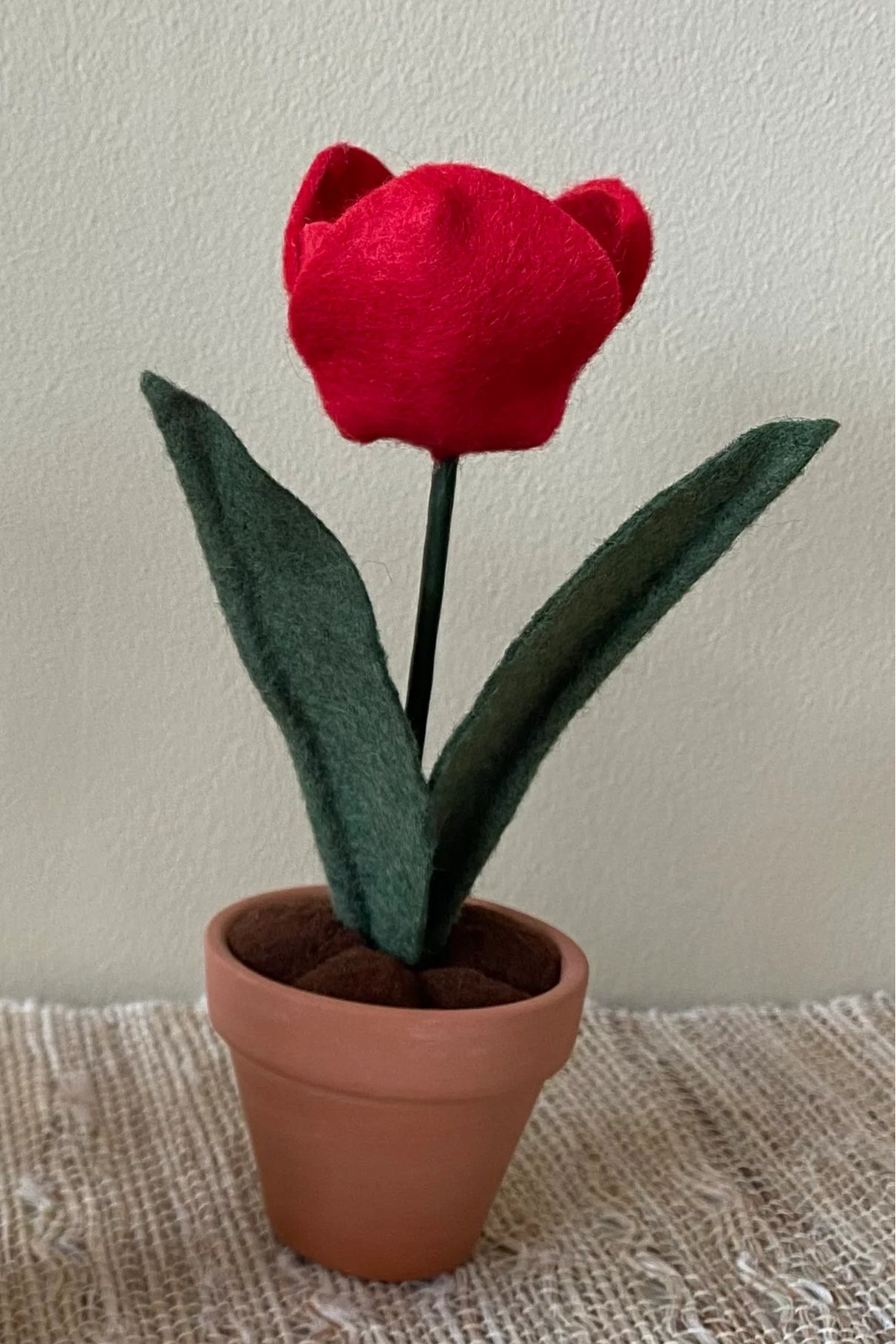 The Handmade Felt Tulip by ForgetBKnot is shown on a piece of fabric against a beige background.