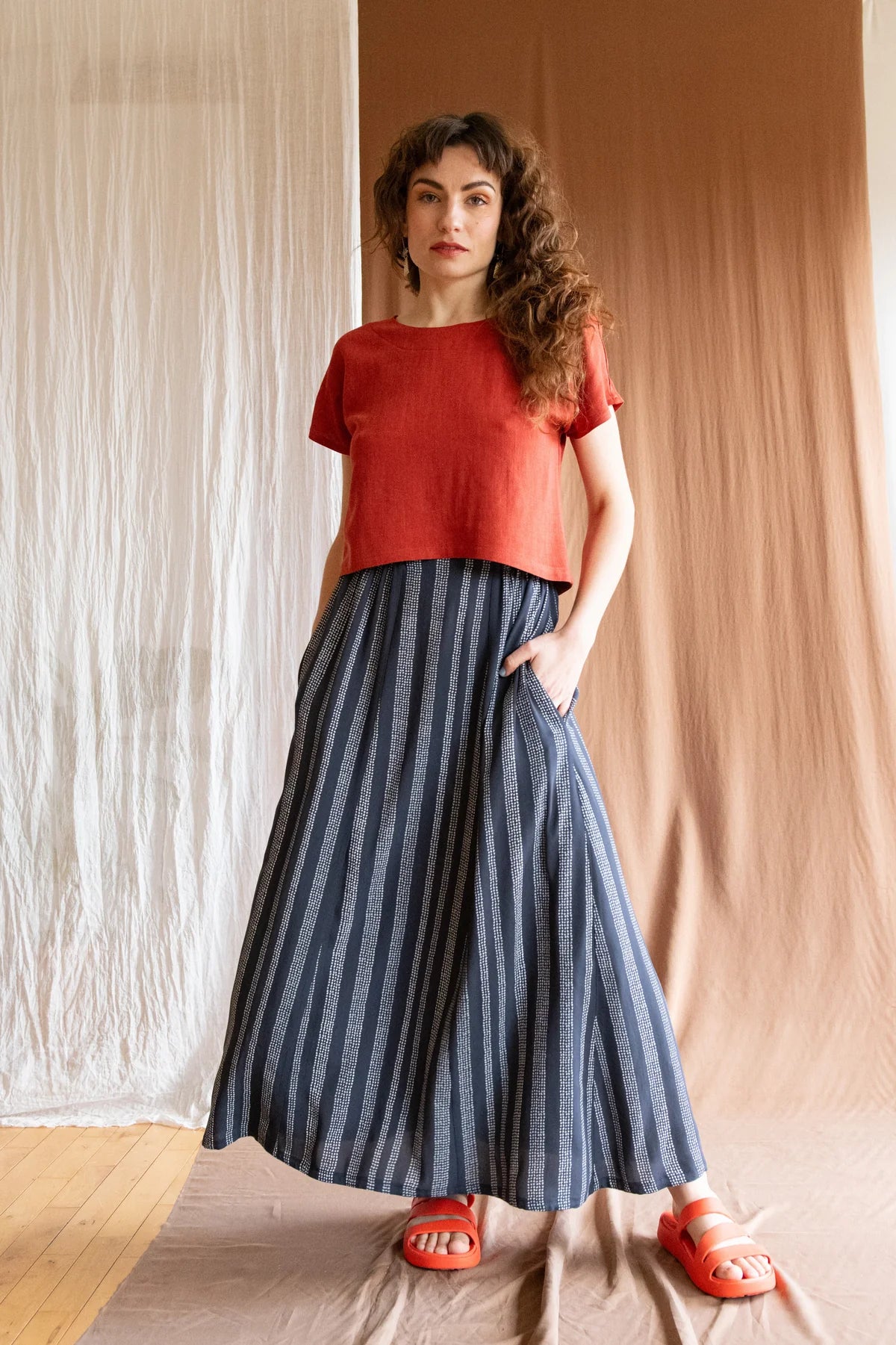 A woman wearing the Brendola Top by Kazak in Rust, a reversible top with a v-neck and snap front on one side and a round neck on the other side. It has short extended sleeves and a relaxed, cropped fit. She is wearing it with the Verona Skirt in Charcoal Dots and standing in front of a brown curtain.