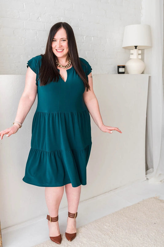 A woman wearing the Fit and Flare Dress by Dotty in Emerald, featuring a double ruffled short sleeve, notched V-neck, and tiered knee-length skirt. She is standing in front of a white wall.