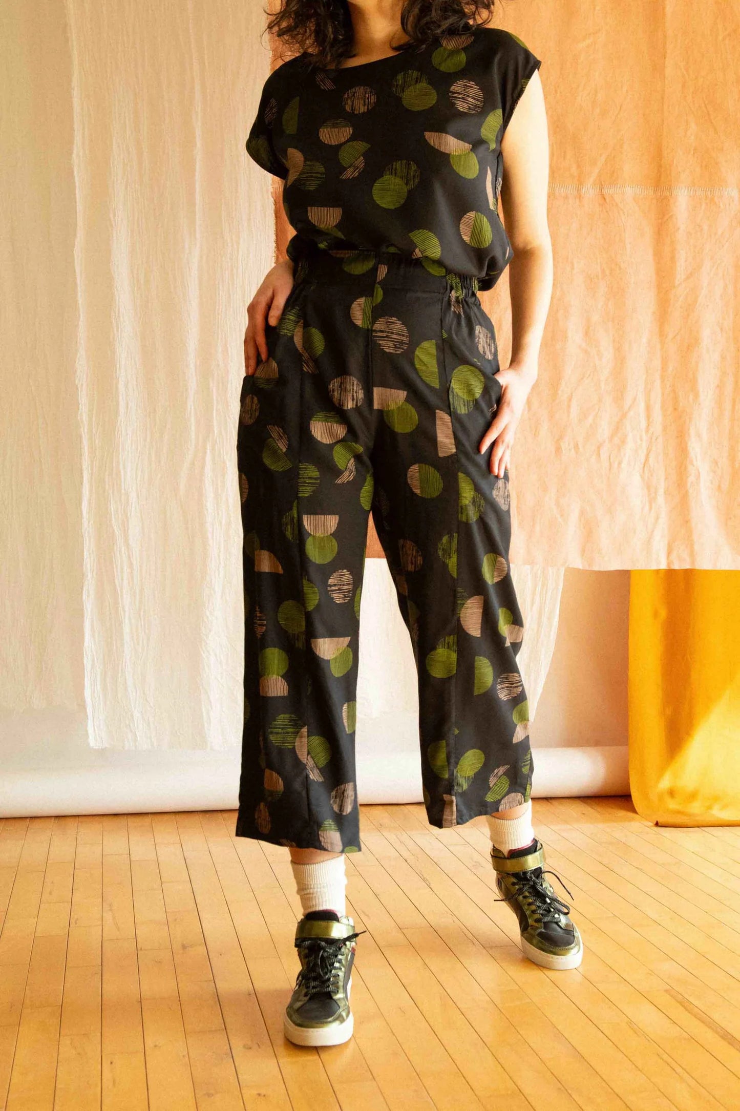 Neck down view of a woman wearing the Sandrigo Top by Kazak in Green Moon, with short extended sleeves, a round and wide neck, and a loose fit. She is wearing it with matching pants and standing in front of a brown curtain.