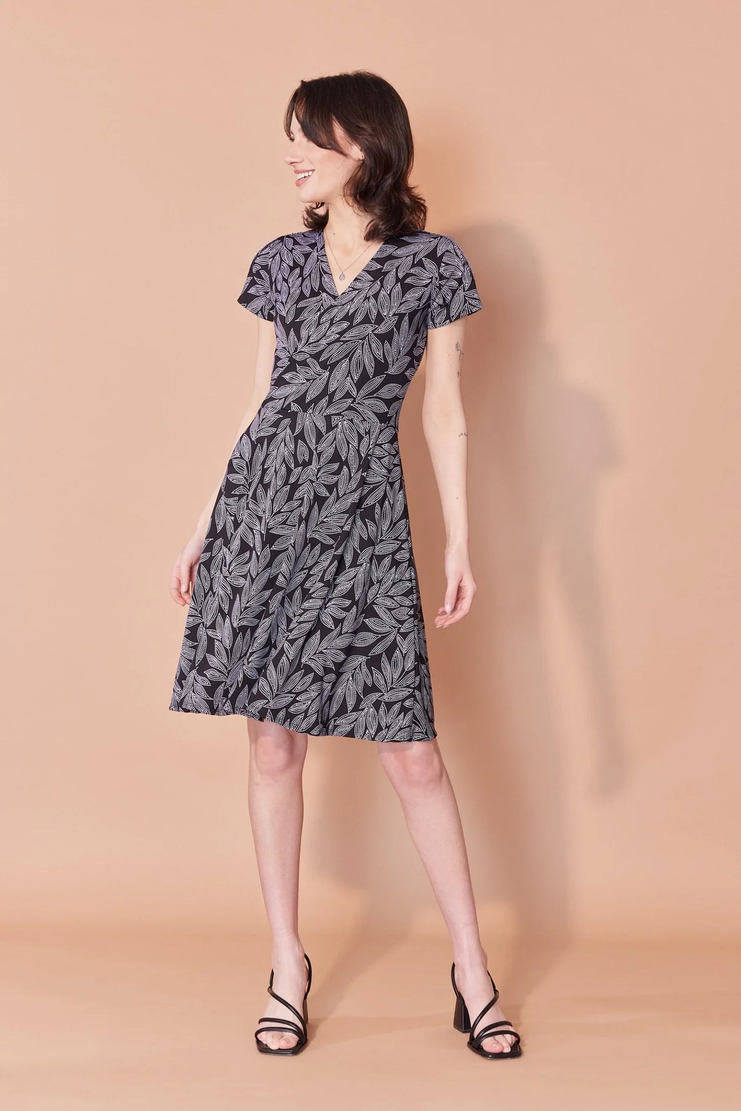 A woman wearing the Josephine Dress by Cherry Bobin in Black and White Leaves, with a wrap-over front, defined waist, short sleeves, and A-line above the knee skirt. She is standing in front of a beige background.