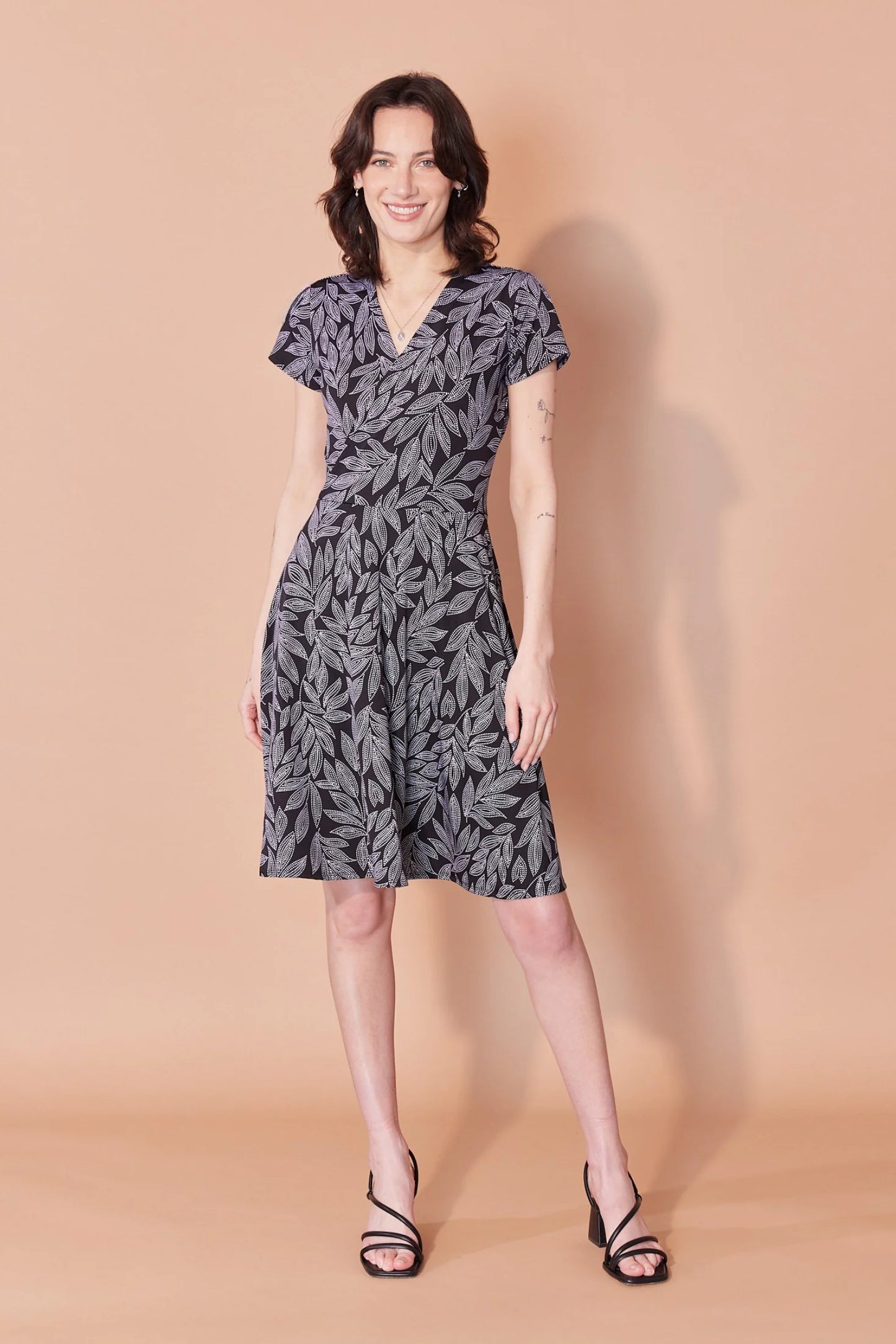 A woman wearing the Josephine Dress by Cherry Bobin in Black and White Leaves, with a wrap-over front, defined waist, short sleeves, and A-line above the knee skirt. She is standing in front of a beige background.