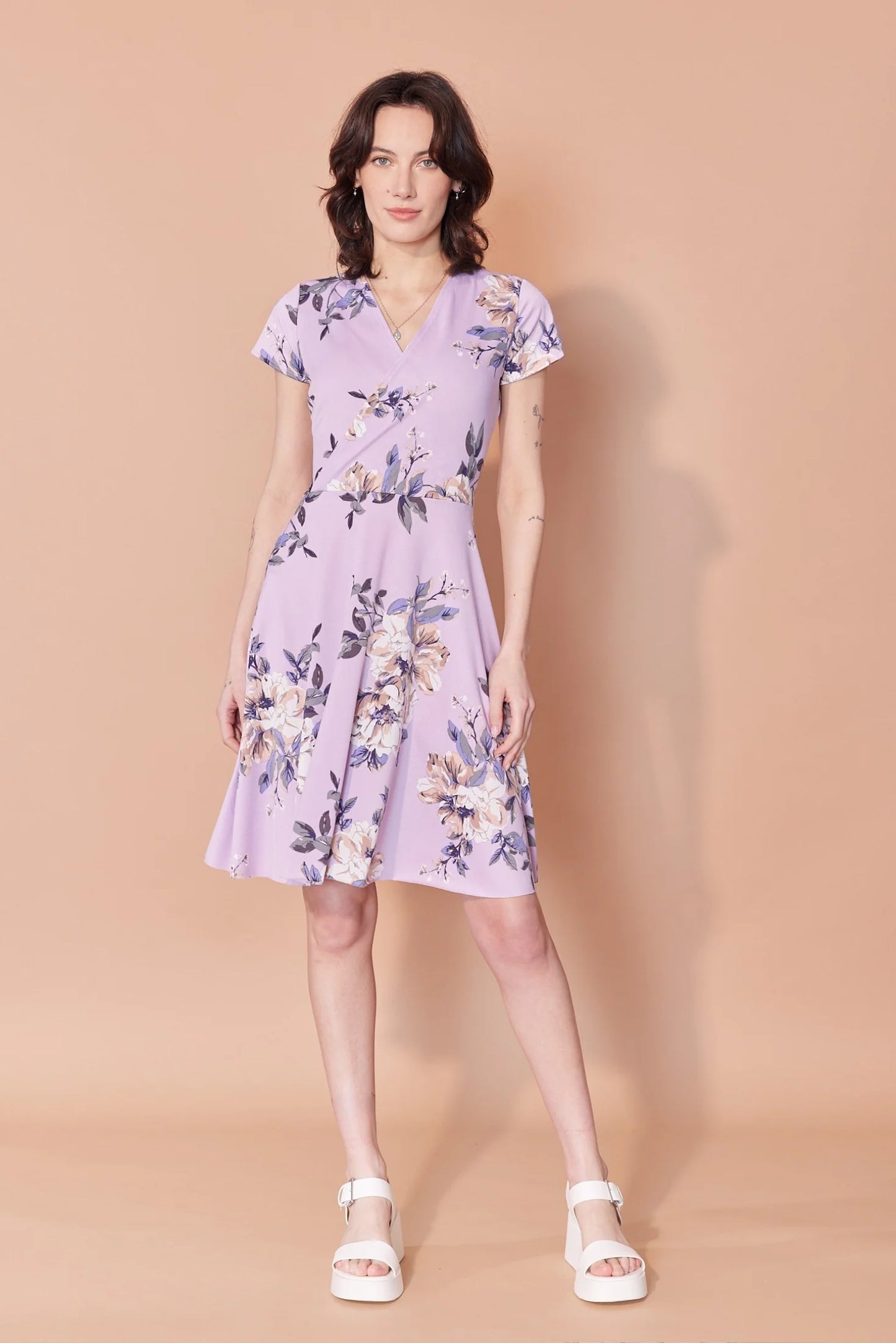 A woman wearing the Josephine Dress by Cherry Bobin in Lilac Floral, with a wrap-over front, defined waist, short sleeves, and A-line above the knee skirt. She is standing in front of a beige background.
