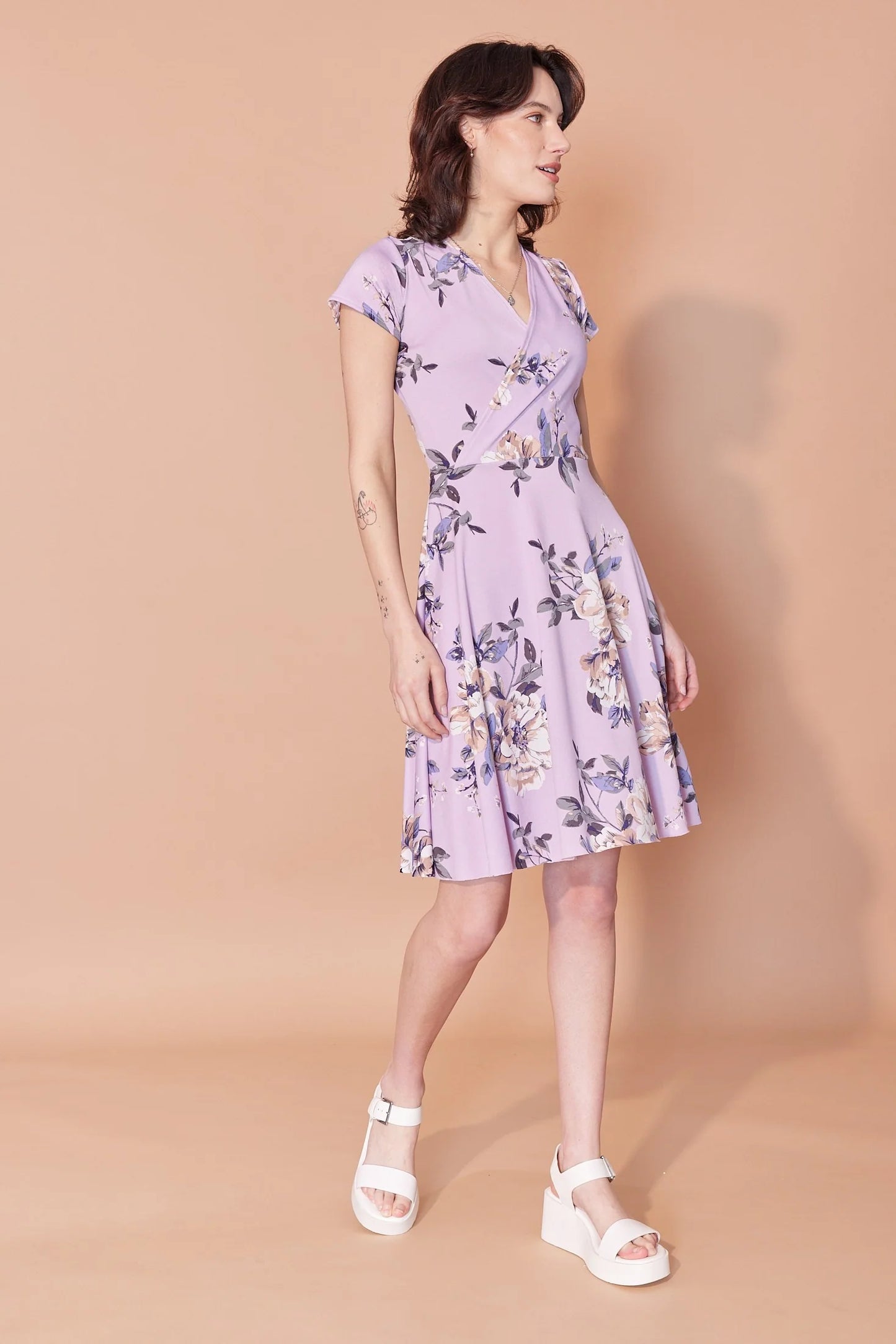 A woman wearing the Josephine Dress by Cherry Bobin in Lilac Floral, with a wrap-over front, defined waist, short sleeves, and A-line above the knee skirt. She is standing in front of a beige background.