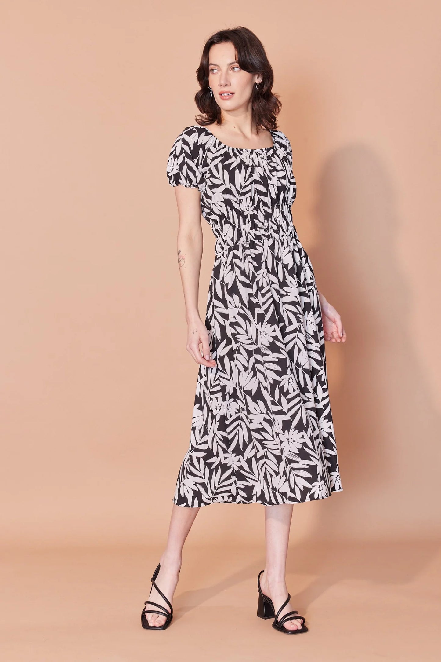 A woman wearing the Victoria Dress by Cherry Bobin in Black and Ivory Leaves print, featuring a scooped peasant neckline, short gathered sleeves, an empire waist with ruching and elastic, and a midi-length skirt. She is standing in front of a beige background.