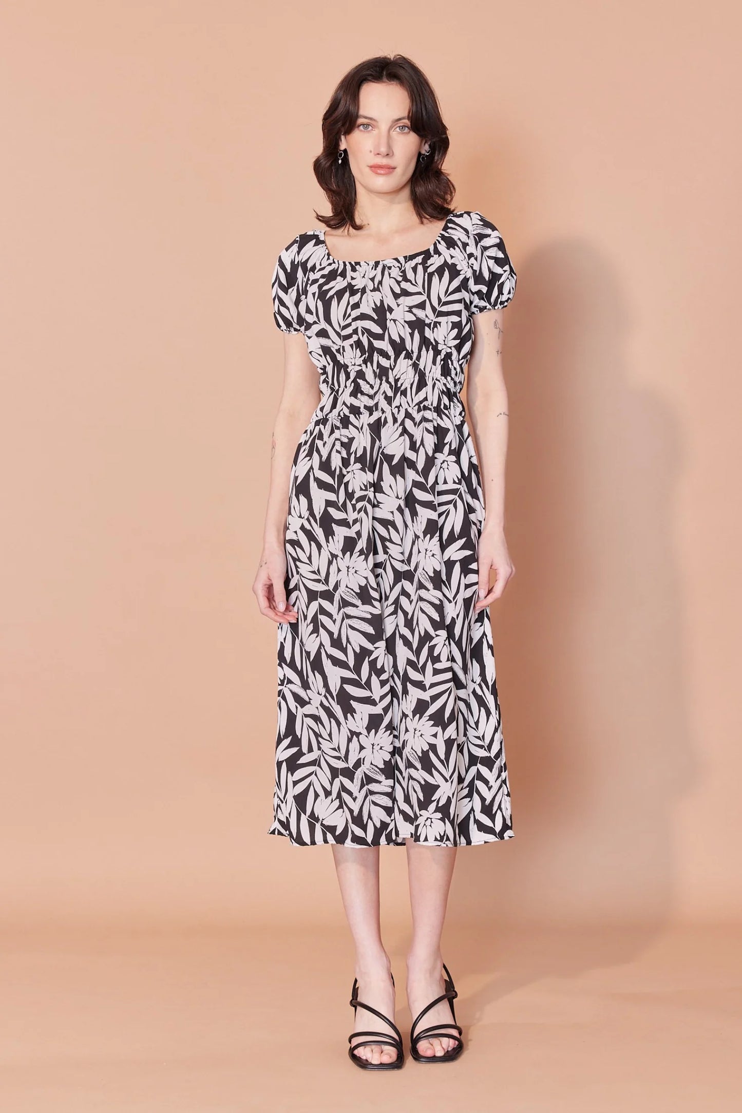 A woman wearing the Victoria Dress by Cherry Bobin in Black and Ivory Leaves print, featuring a scooped peasant neckline, short gathered sleeves, an empire waist with ruching and elastic, and a midi-length skirt. She is standing in front of a beige background.