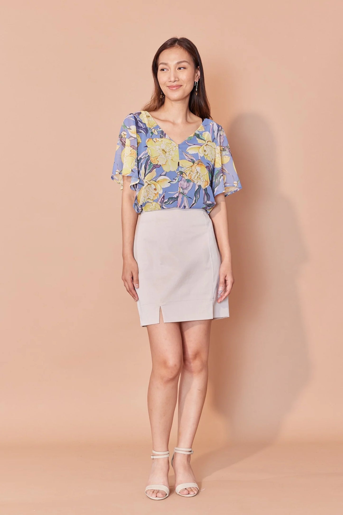 A woman wearing the Anastasia Top by Cherry Bobin in Yellow Peony print, featuring a v-neck, full elbow-length sleeves, and an open back with tie closures. She is wearing it with a white skirt and standing in front of a beige background.