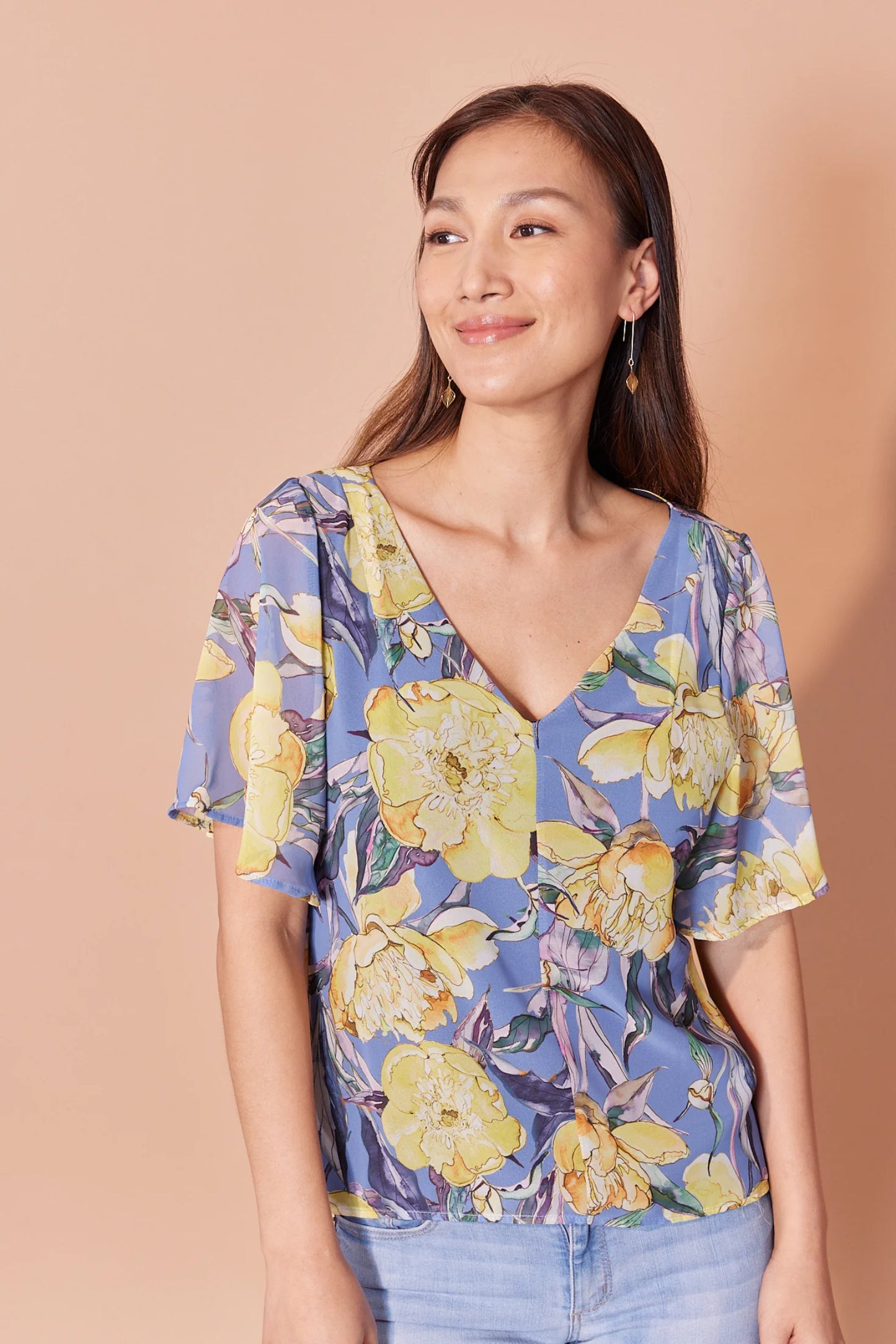 A woman wearing the Anastasia Top by Cherry Bobin in Yellow Peony print, eaturing a v-neck, full elbow-length sleeves, and an open back with tie closures. She is wearing it with jeans and standing in front of a beige background.