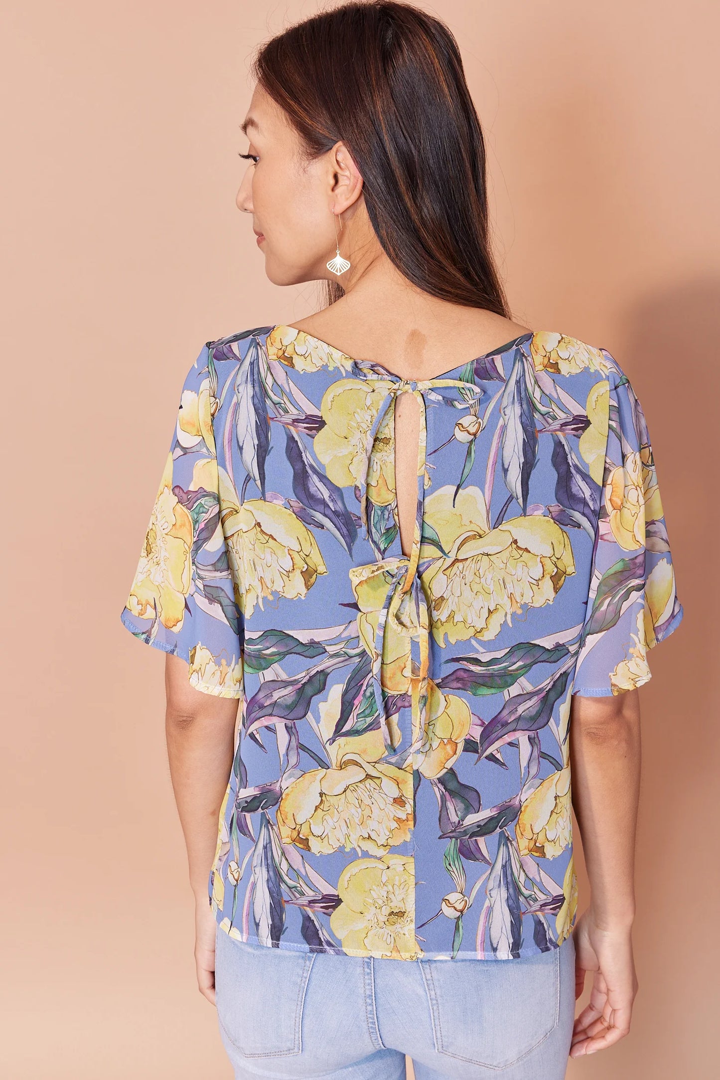Back waist-up view of woman wearing the Anastasia Top by Cherry Bobin in Yellow Peony print, eaturing a v-neck, full elbow-length sleeves, and an open back with tie closures. She is wearing it with jeans and standing in front of a beige background.