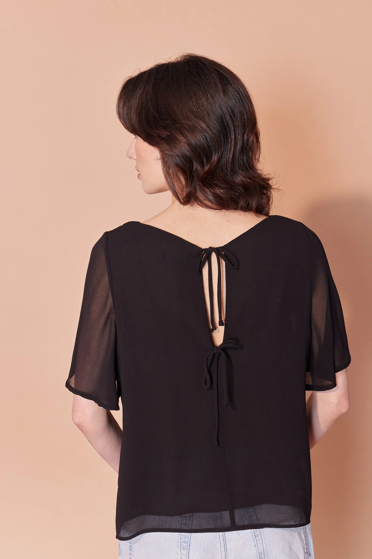 Back waist-up view of a woman wearing the Anastasia Top by Cherry Bobin in Black, featuring a v-neck, full elbow-length sleeves, and an open back with tie closures. She is wearing it with jeans and standing in front of a beige background.