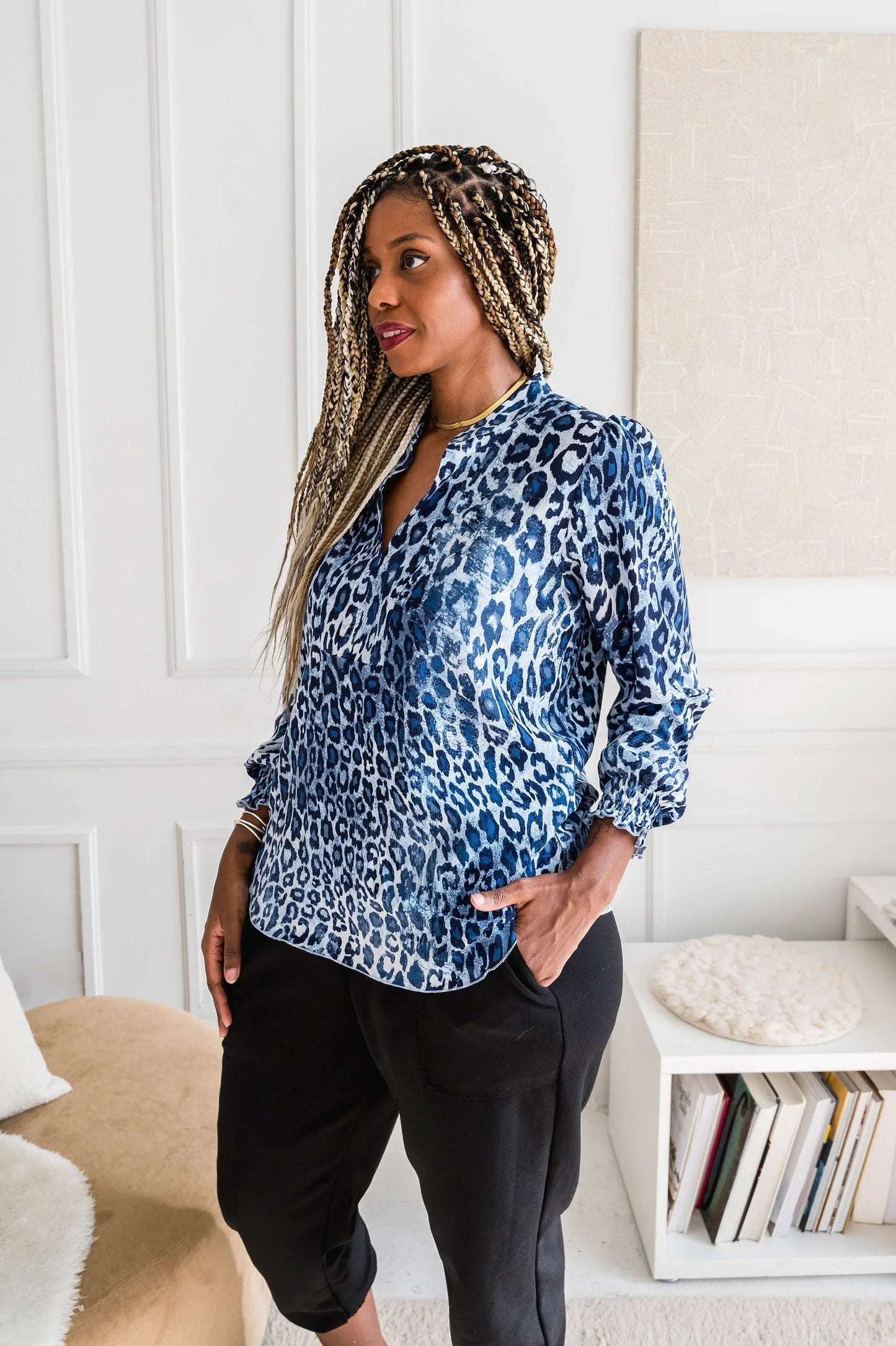 3/4 shot of woman wearing the Blue Leopard Blouse by Dotty, featuring a V-neck, banded collar, and long sleeves with smocked cuffs. It has a flowy fit and and rounded hem. She is wearing it with black pants and standing in front of a white wall.