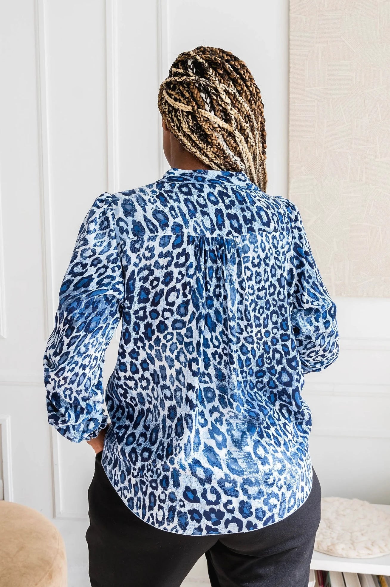 Back 3/4 view of a woman wearing the Blue Leopard Blouse by Dotty, featuring a V-neck, banded collar, and long sleeves with smocked cuffs. It has a flowy fit and and rounded hem. She is wearing it with black pants and standing in front of a white wall.
