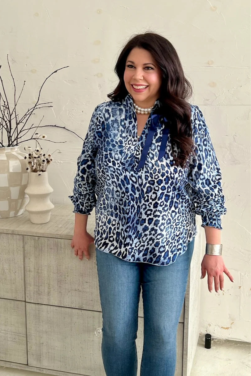 A woman wearing the Blue Leopard Blouse by Dotty, featuring a V-neck, banded collar, and long sleeves with smocked cuffs. It has a flowy fit and and rounded hem. She is wearing it with jeans and standing in front of a beige sideboard.