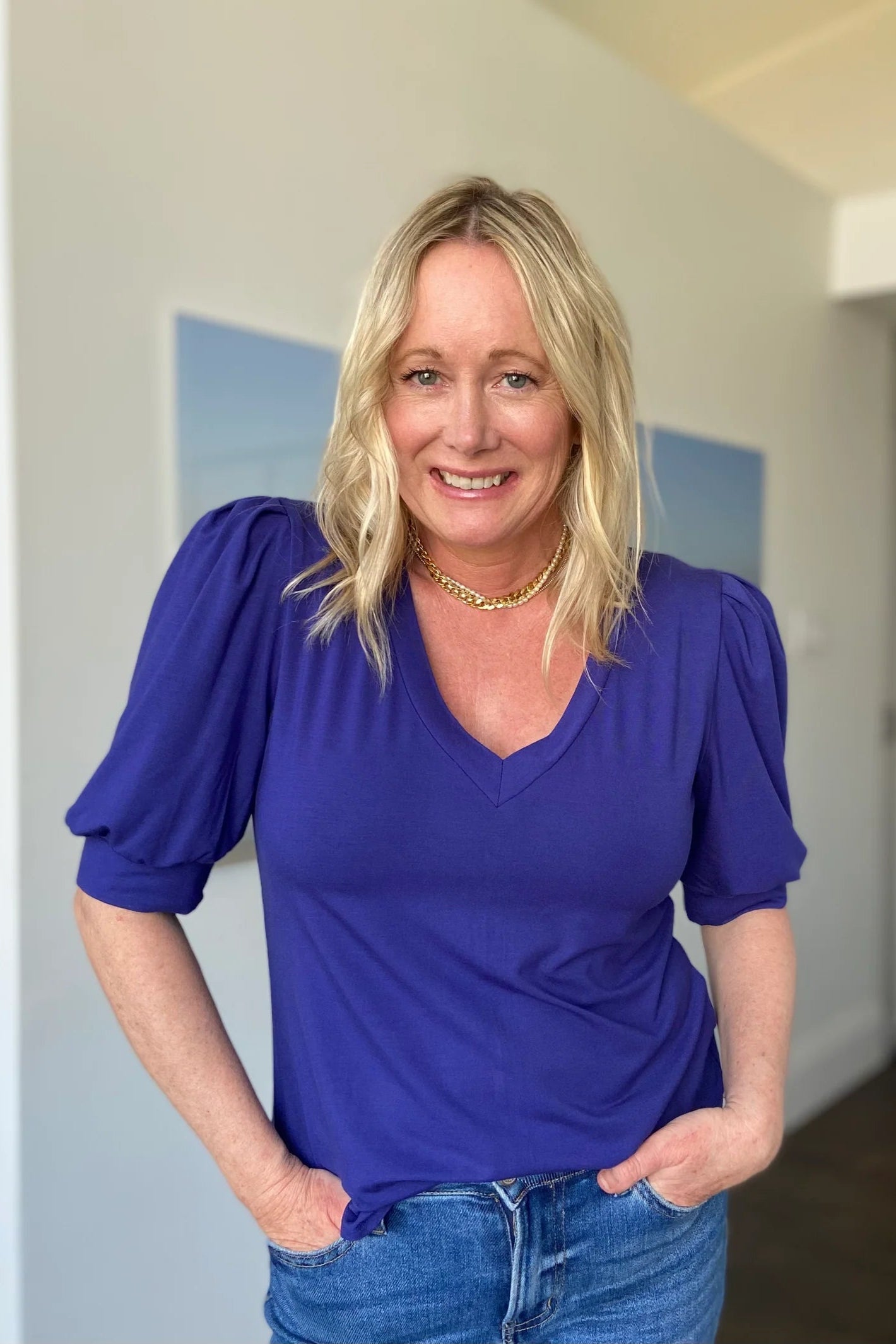 A woman wearing the Darling Tee by Dotty in Blueberry, with a v-neck, pleats at the shoulders, elbow-length sleeves, and a rounded hem. She is wearing it with jeans and is standing in front of a white wall.