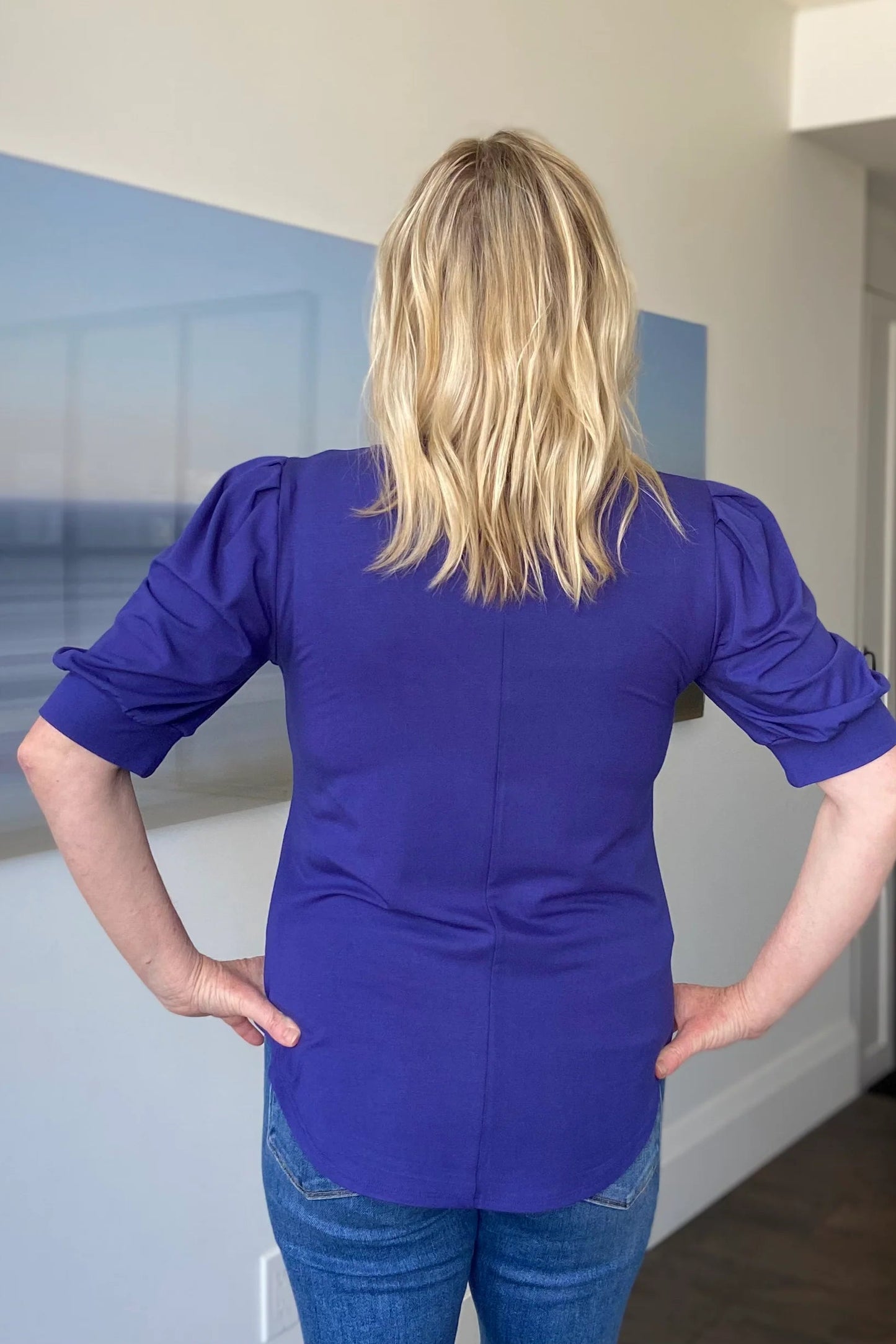 Back view of a woman wearing the Darling Tee by Dotty in Blueberry, with a v-neck, pleats at the shoulders, elbow-length sleeves, and a rounded hem. She is wearing it with jeans and is standing in front of a white wall.