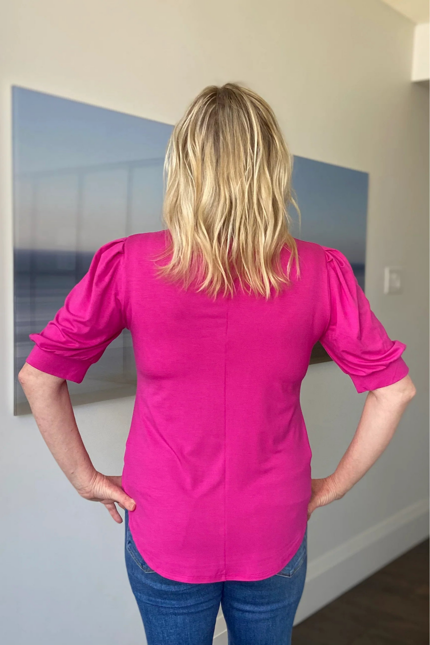 Back view of a woman wearing the Darling Tee by Dotty in Hot Pink, with a v-neck, pleats at the shoulders, elbow-length sleeves, and a rounded hem. She is wearing it with jeans and is standing in front of a white wall.
