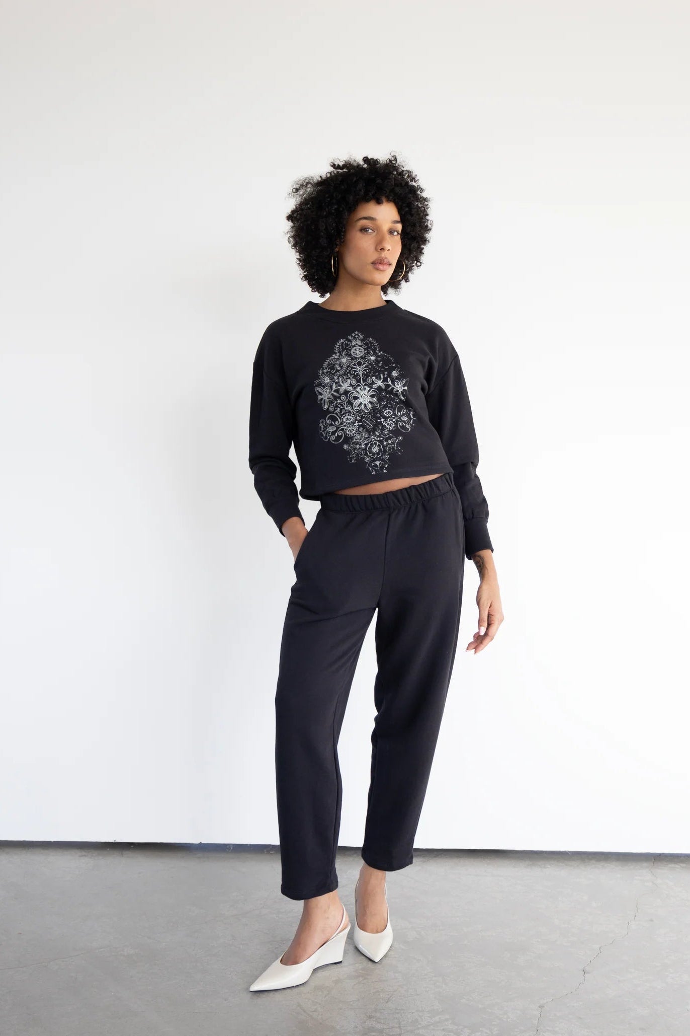 A woman wearing the Baxter Fleece Pants by Eve Lavoie, ankle-length joggers with an elastic waist and side pockets. She is wearing them with the Hanay Sweatshirt in Black and standing in front of a white background.