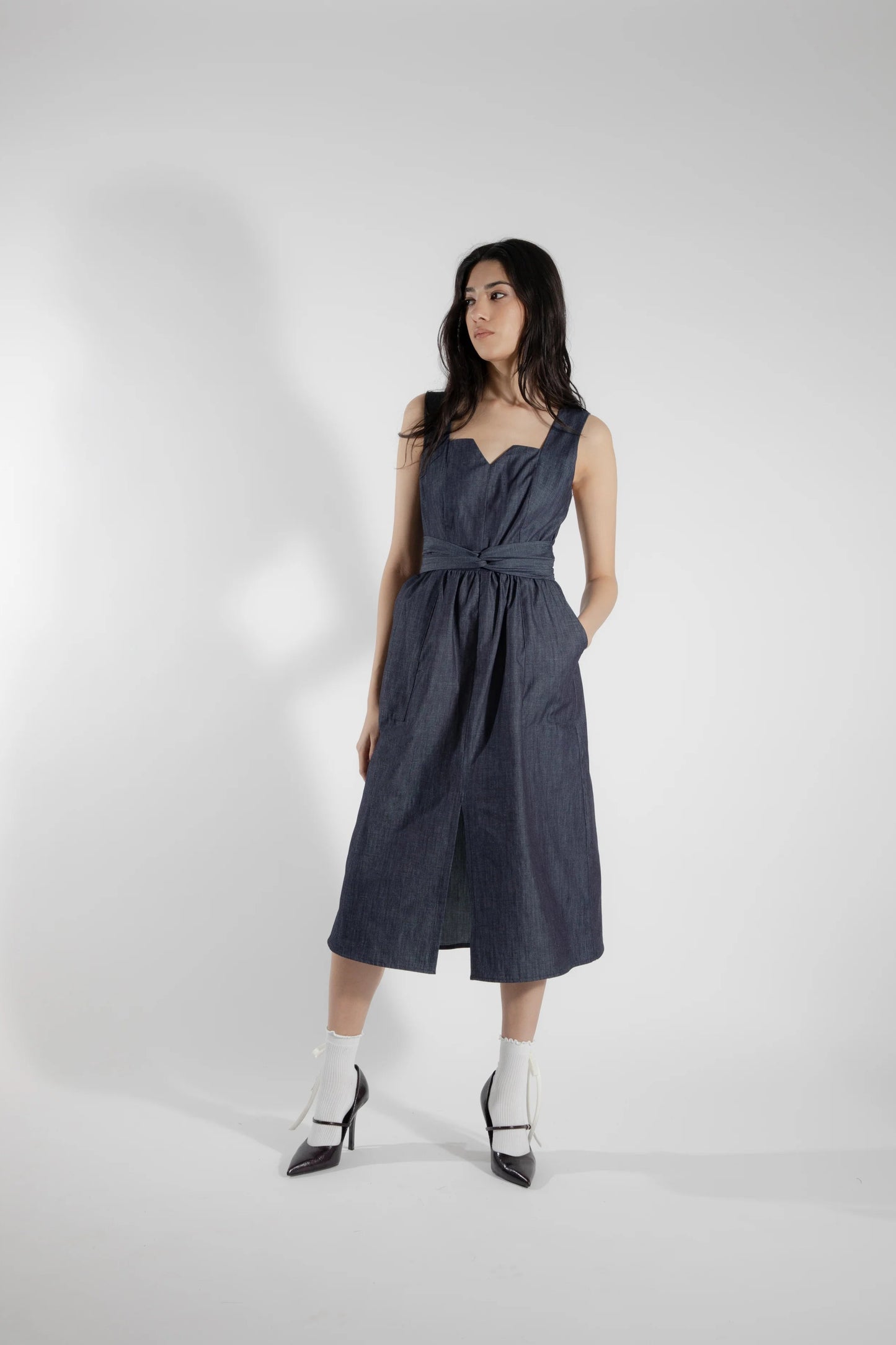 A woman wearing the Cornifle Dress by Eve Lavoie in Denim, a sleeveless dress with a fitted bodice, wide straps, square neckline with a V-notch, full skirt that falls below the knee, a front slit, and patch pockets. She is standing in front of a white background.