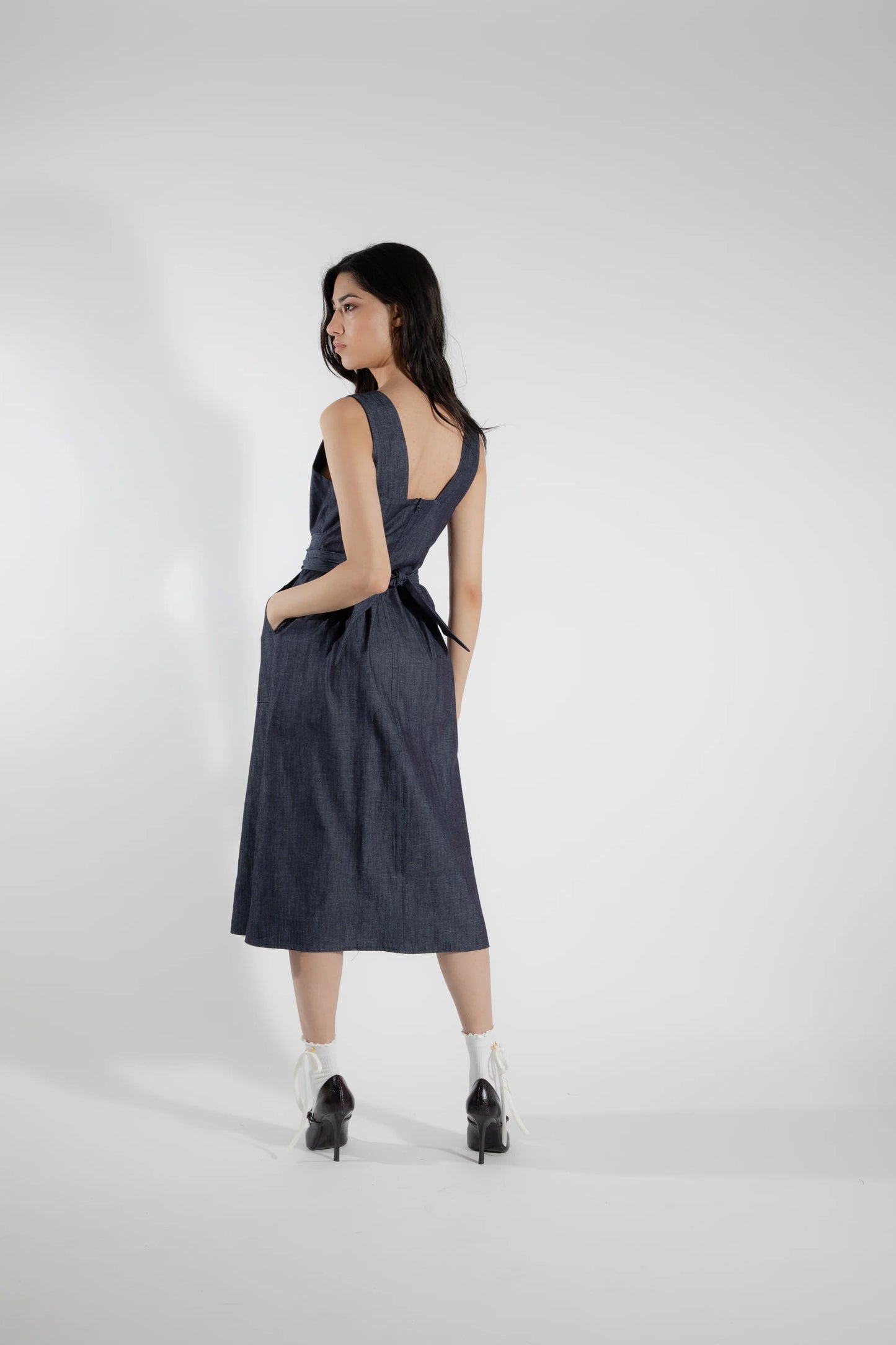 Back view of a woman wearing the Cornifle Dress by Eve Lavoie in Denim, a sleeveless dress with a fitted bodice, wide straps, square neckline with a V-notch, full skirt that falls below the knee, a front slit, and patch pockets. She is standing in front of a white background.
