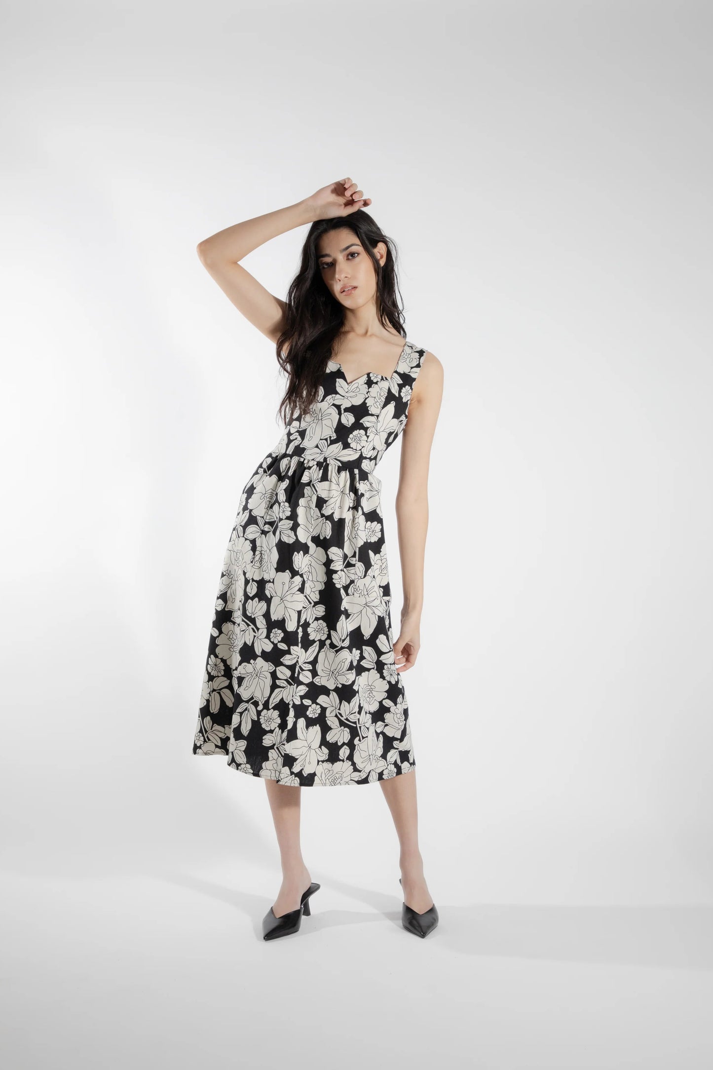 A woman wearing the Cornifle Dress by Eve Lavoie in black and white Floral, a sleeveless dress with a fitted bodice, wide straps, square neckline with a V-notch, full skirt that falls below the knee, a front slit, and patch pockets. She is standing in front of a white background.