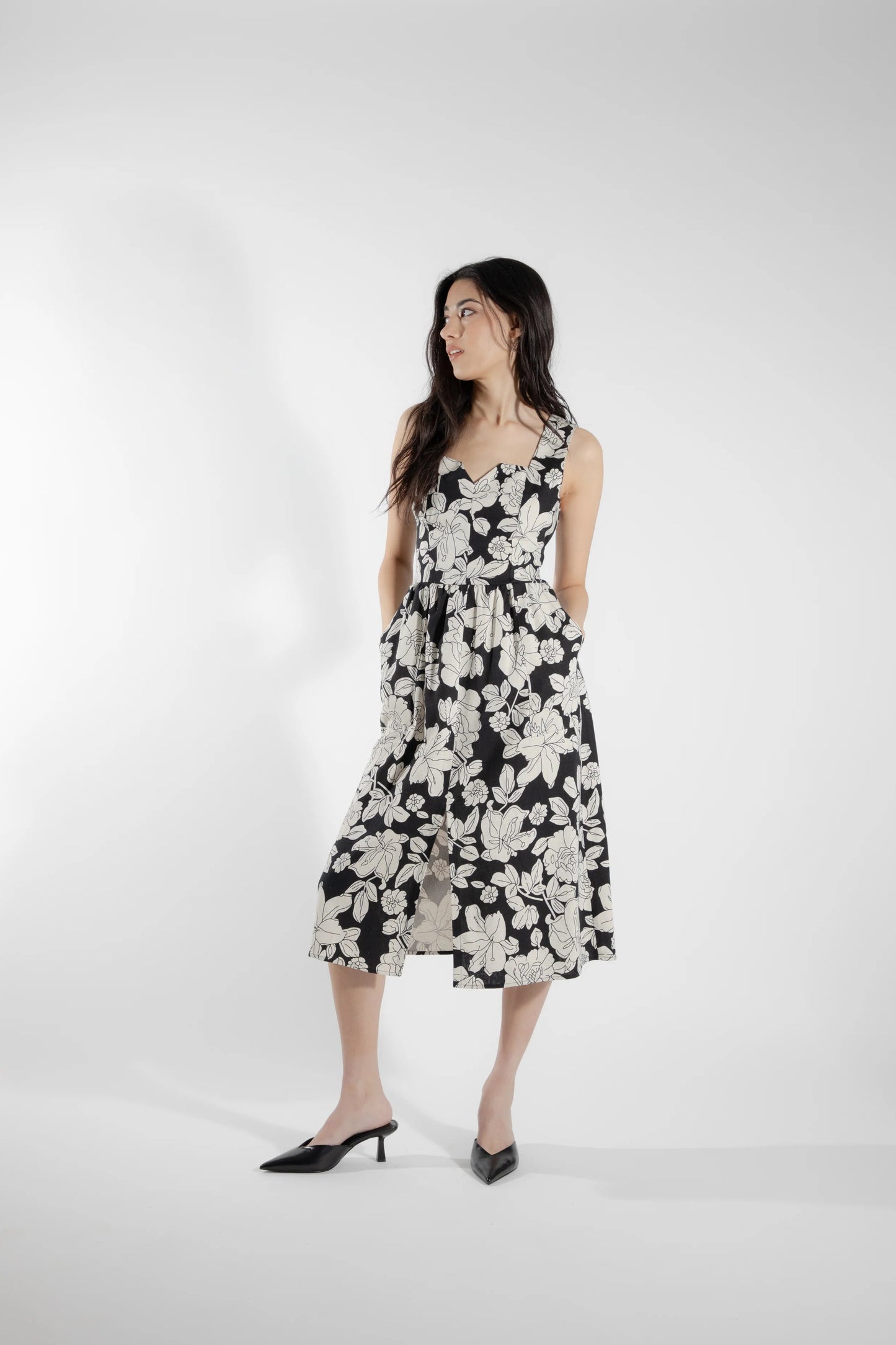 A woman wearing the Cornifle Dress by Eve Lavoie in black and white Floral, a sleeveless dress with a fitted bodice, wide straps, square neckline with a V-notch, full skirt that falls below the knee, a front slit, and patch pockets. She is standing in front of a white background.