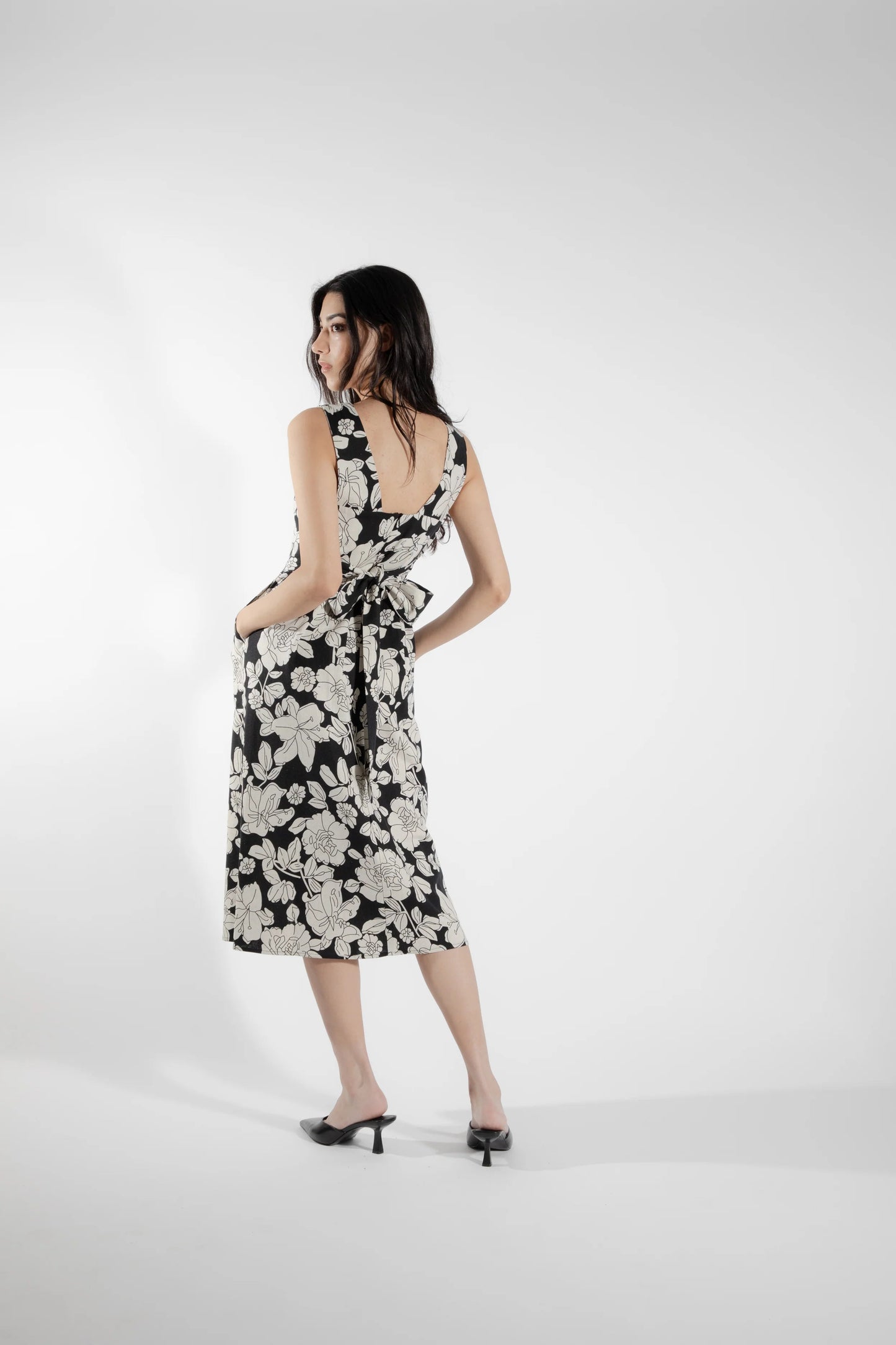 Back view of a woman wearing the Cornifle Dress by Eve Lavoie in black and white Floral, a sleeveless dress with a fitted bodice, wide straps, square neckline with a V-notch, full skirt that falls below the knee, a front slit, and patch pockets. She is standing in front of a white background.