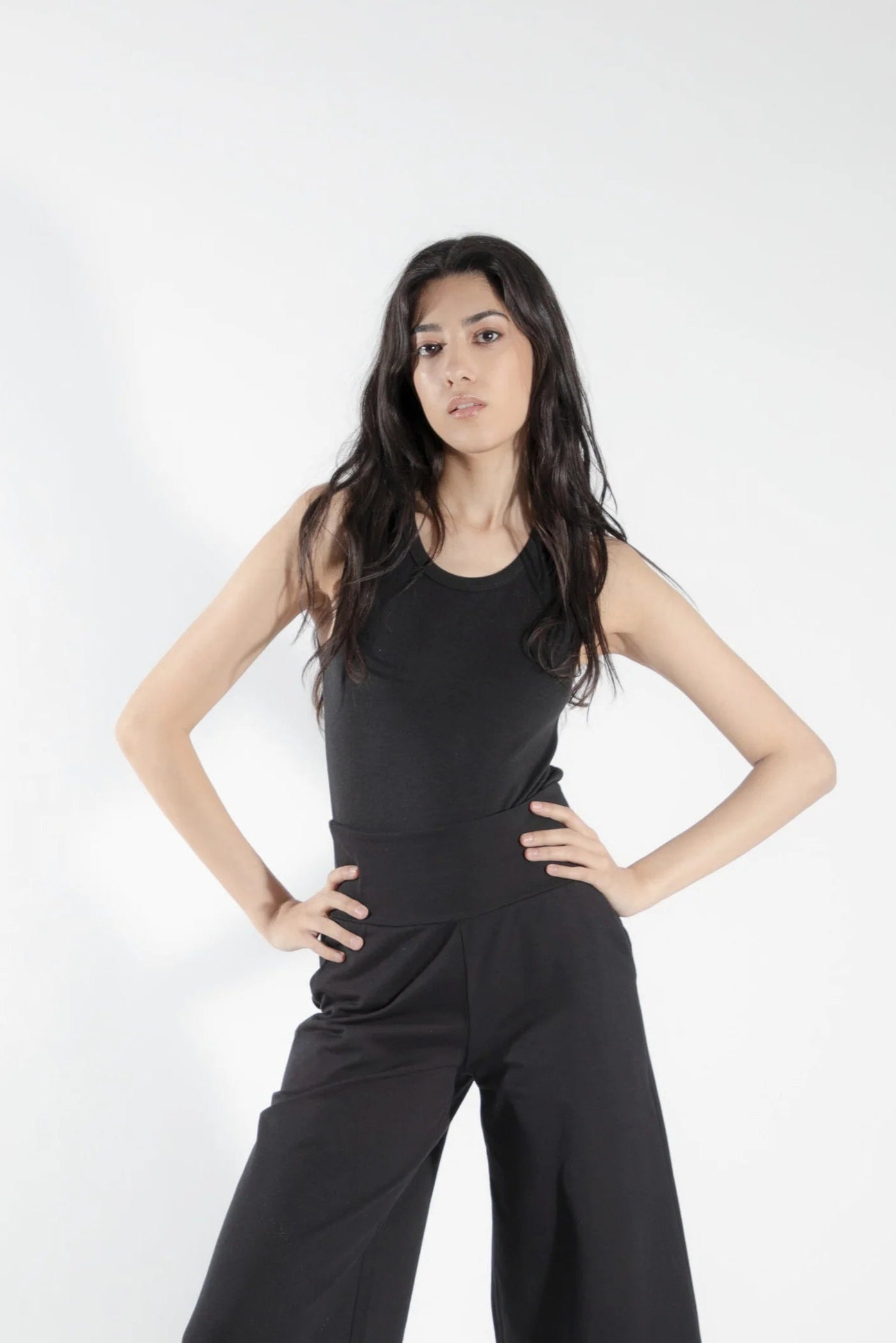 A woman wearing the Rubanier Camisole by Eve Lavoie in Black, featuring wide straps, a round neck, and a slim fit. She is wearing it with the Chara Pants in Black and is standing in front of a white background.