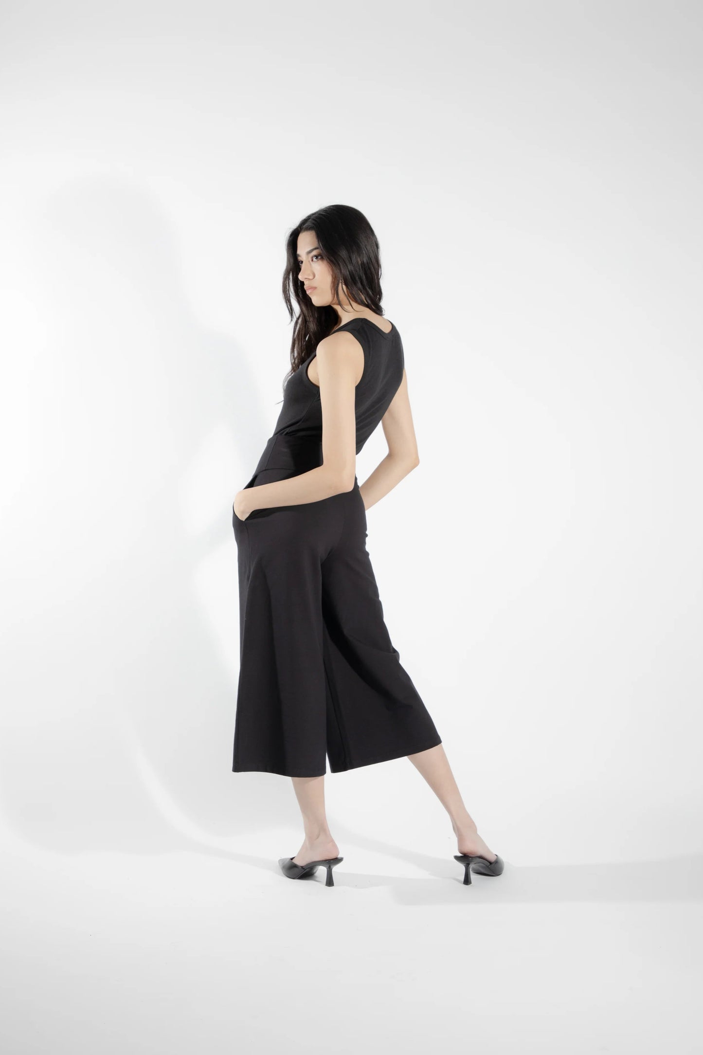 Back view of a woman wearing the Rubanier Camisole by Eve Lavoie in Black, featuring wide straps, a round neck, and a slim fit. She is wearing it with the Chara Pants in Black and is standing in front of a white background.