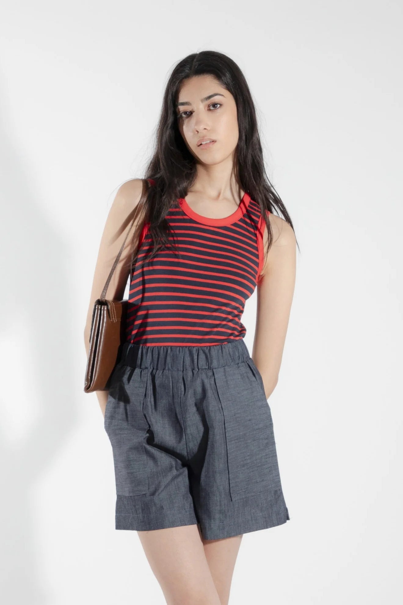 A woman wearing the Rubanier Camisole by Eve Lavoie in Red Stripe, featuring wide straps, a round neck, contrast piping at the collar and arm holes, and a slim fit. She is wearing it with denim shorts and is standing in front of a white background.