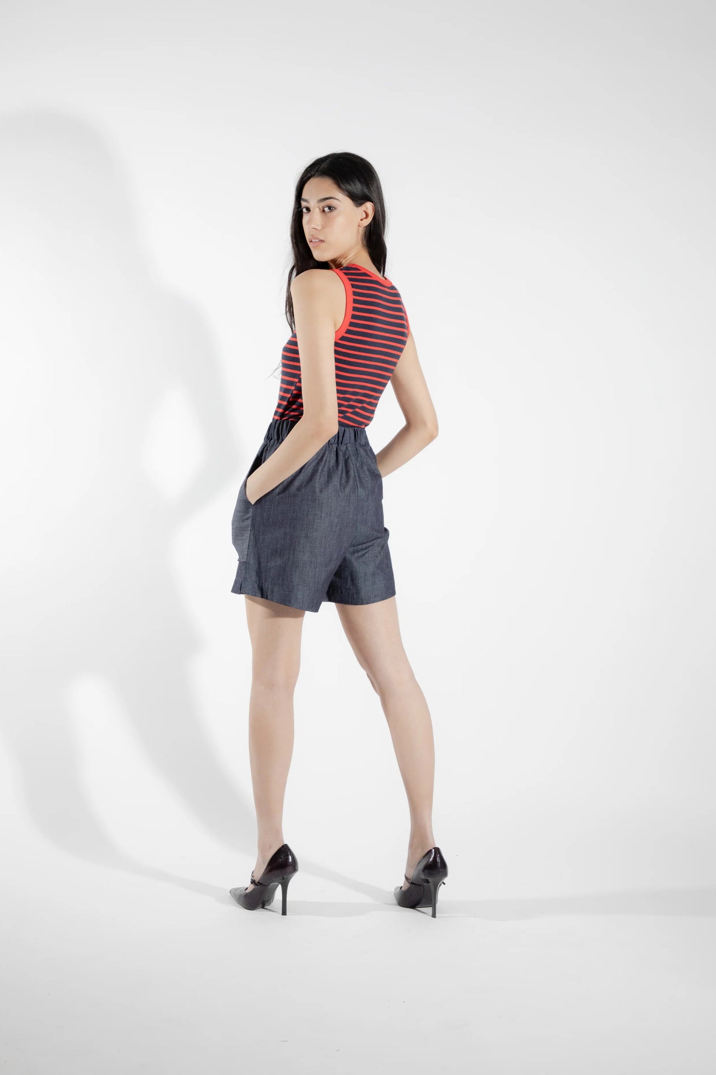 Back view of a woman wearing the Rubanier Camisole by Eve Lavoie in Red Stripe, featuring wide straps, a round neck, contrast piping at the collar and arm holes, and a slim fit. She is wearing it with denim shorts and is standing in front of a white background.