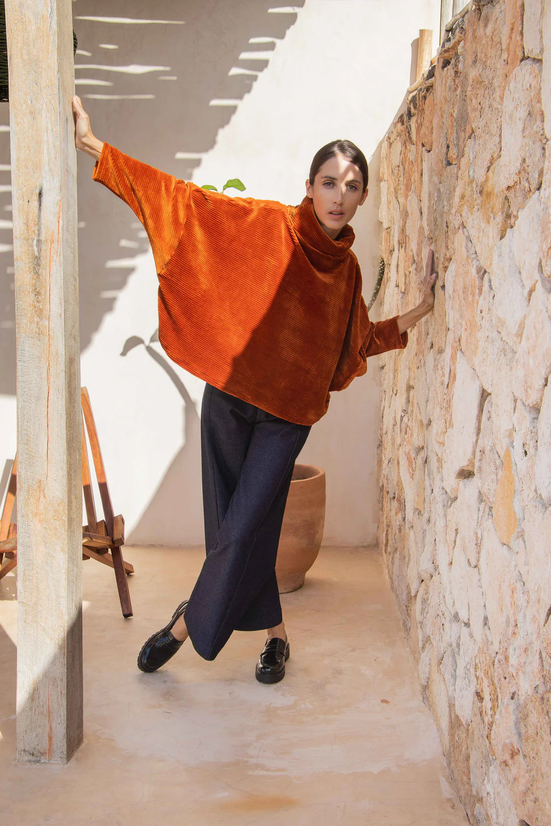 A woman wearing the Vienna Pants by MAS in Jeans, high waisted, wide-legged pants with a zipper and tab. She is wearing them with an orange top and standing in front of a stone wall.