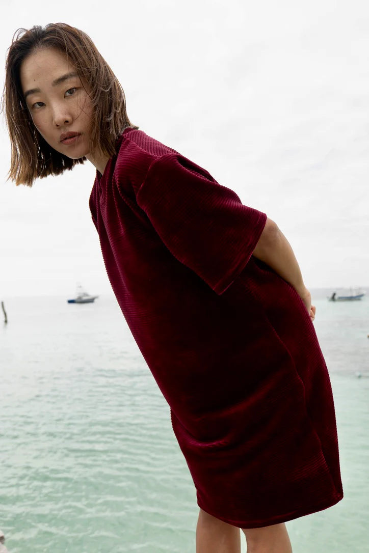 A woman wearing the Mykonos Dress by MAS in Rosewood, a short sleeved boxy, knee-length dress in ribbed velvet fabric. She is standing in front a body of water.