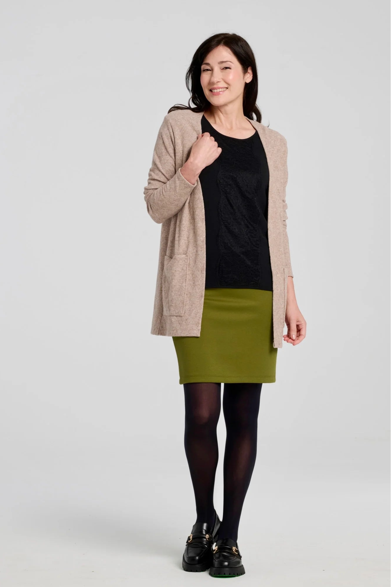 A woman wearing the Veneto Skirt by Kollontai in Granny Smith, a pencil skirt with a wide-pull on waistband that hits above the knee. She is wearing it with a black top and beige cardigan and standing in front of a grey background.