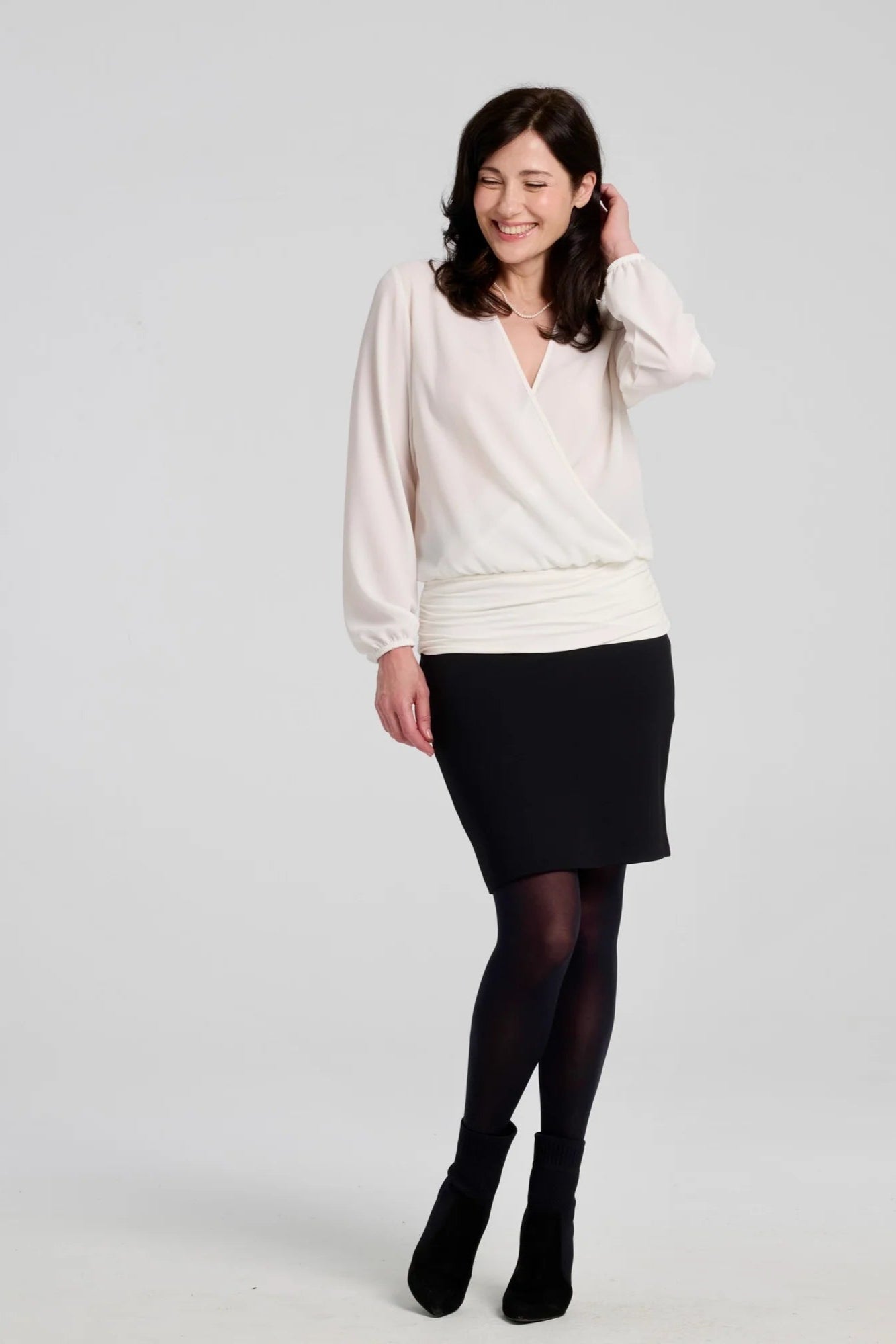 A woman wearing the Veneto Skirt by Kollontai in Black, a pencil skirt with a wide-pull on waistband, back slit, and hem that hits above the knee. She is wearing it with an ivory blouse and standing in front of a grey background.
