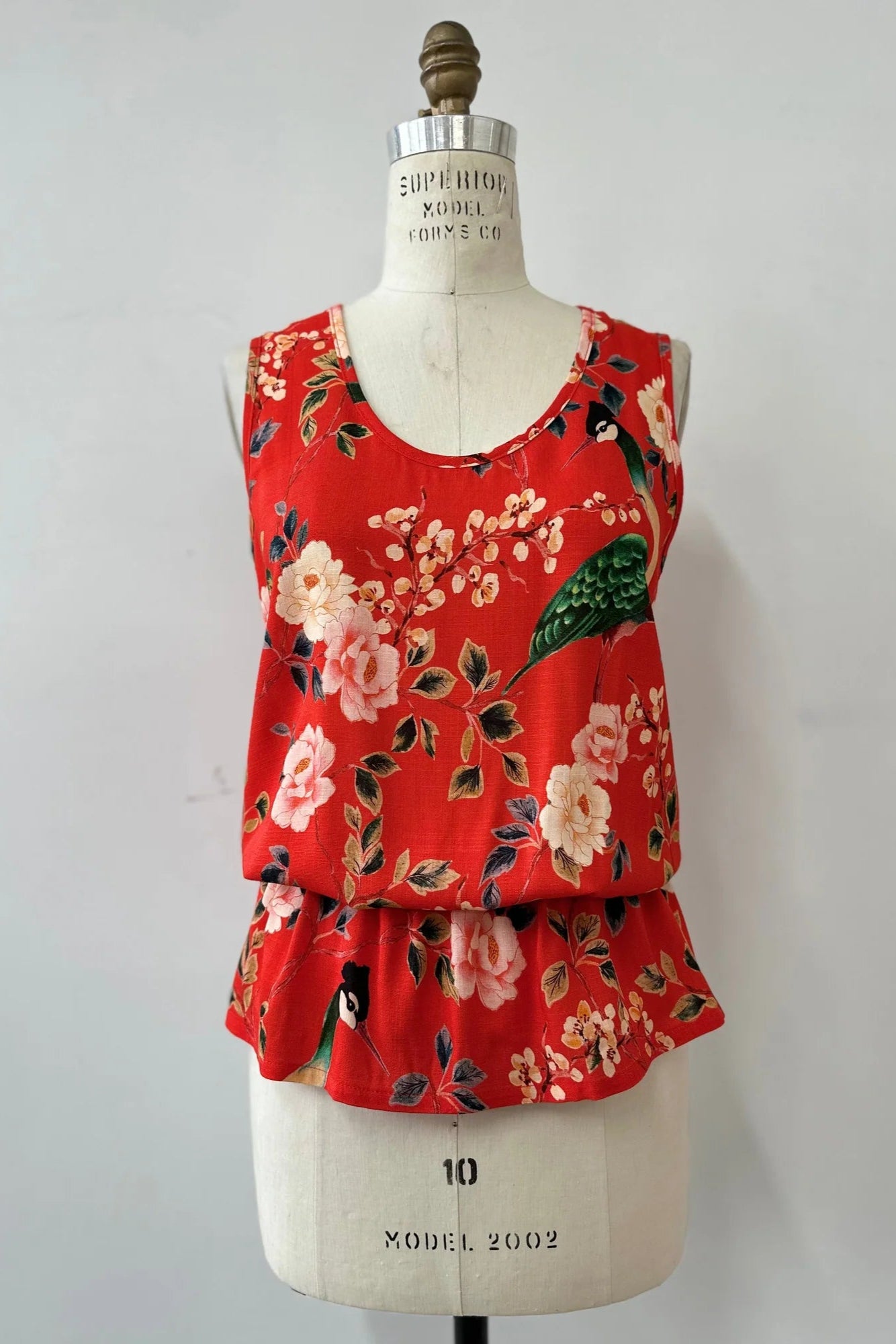 The Yvaine Top by Kollontai in Red, a sleeveless top with a scoop neck, wide straps, an elastic waist that creates a bloused effect, is shown on a mannequin in front of a white background.