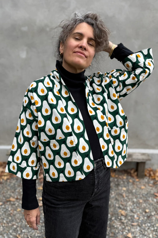 A woman wearing the Magnolia Jacket by Kazak in green and white corduroy Pears print fabric, a short kimono style open jacket with 3/4 sleeves and large patch pockets. She is wearing it with a black top and black jeans and standing in front of a grey wall.