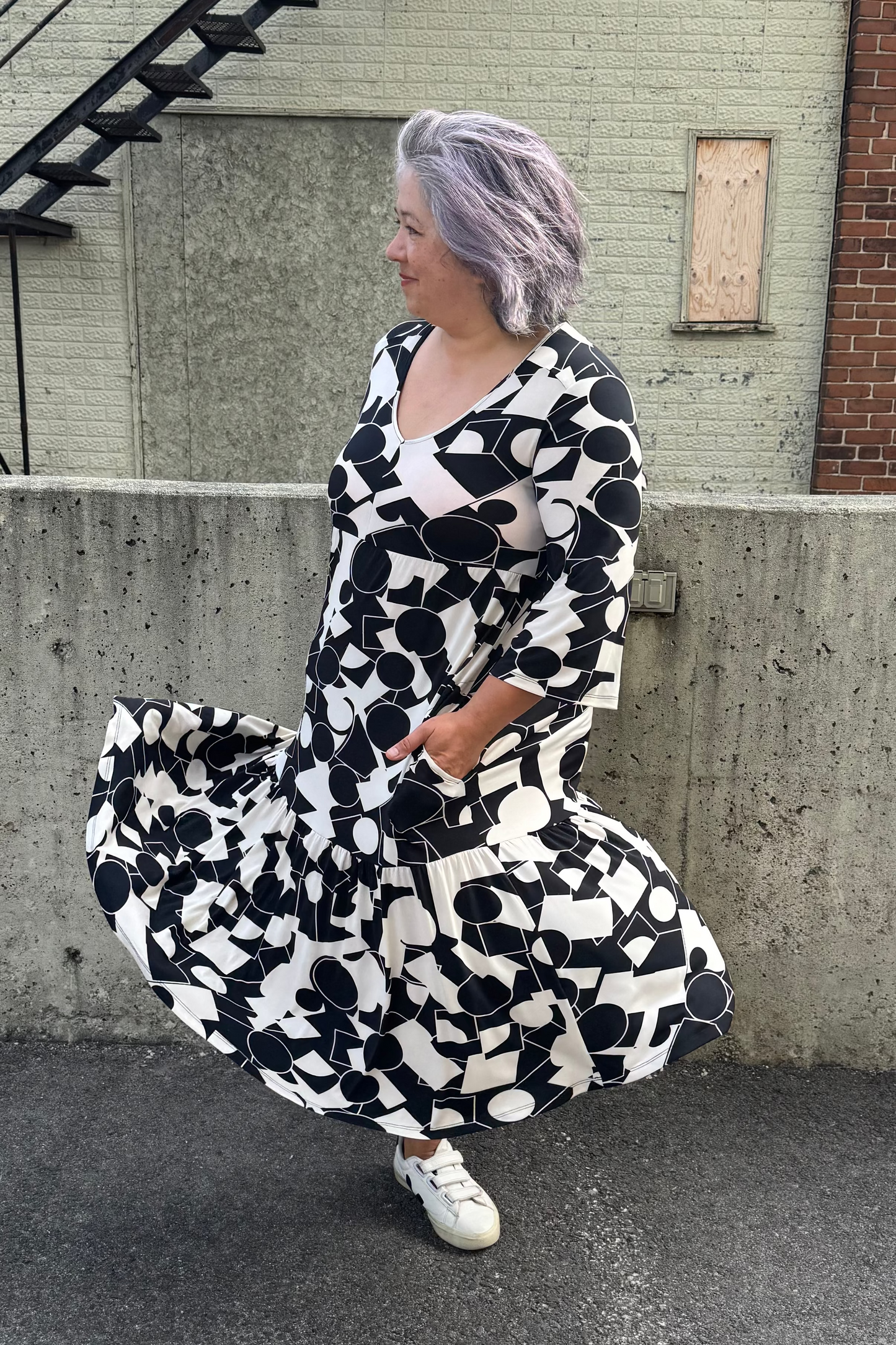 A woman wearing the Farah Dress by Pure Essence in Black/Ivory, a geometric print dress with a v-neck, 3/4-sleeves, baby doll shape, tiered midi length skirt, and front patch pockets. She is twirling in front of a brick building.