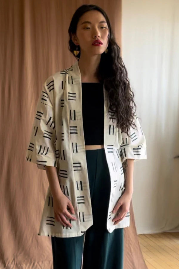 A woman wearing the Mirano Jacket by Kazak in Cream Lines print, an open Kimono-inspired jacket that falls to mid-thigh and has wide 3/4 sleeves. She is wearing it with a black top and green pants and is standing in front of a beige curtain.