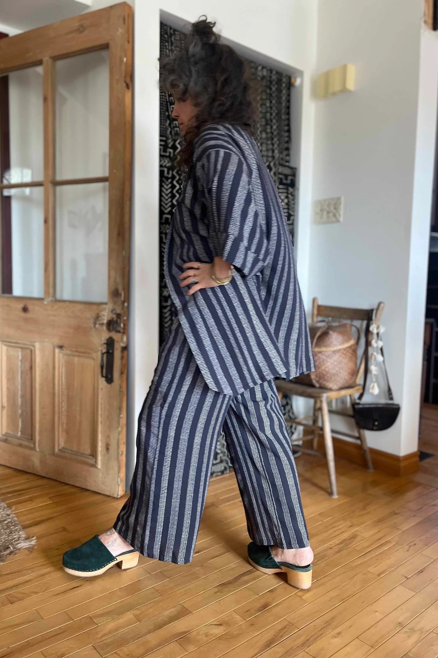 Side view of a woman wearing the Mirano Jacket by Kazak in Charcoal Dots print, an open Kimono-inspired jacket that falls to mid-thigh and has wide 3/4 sleeves. She is wearing it with white top top and matching pants and is standing in front of a wooden door.