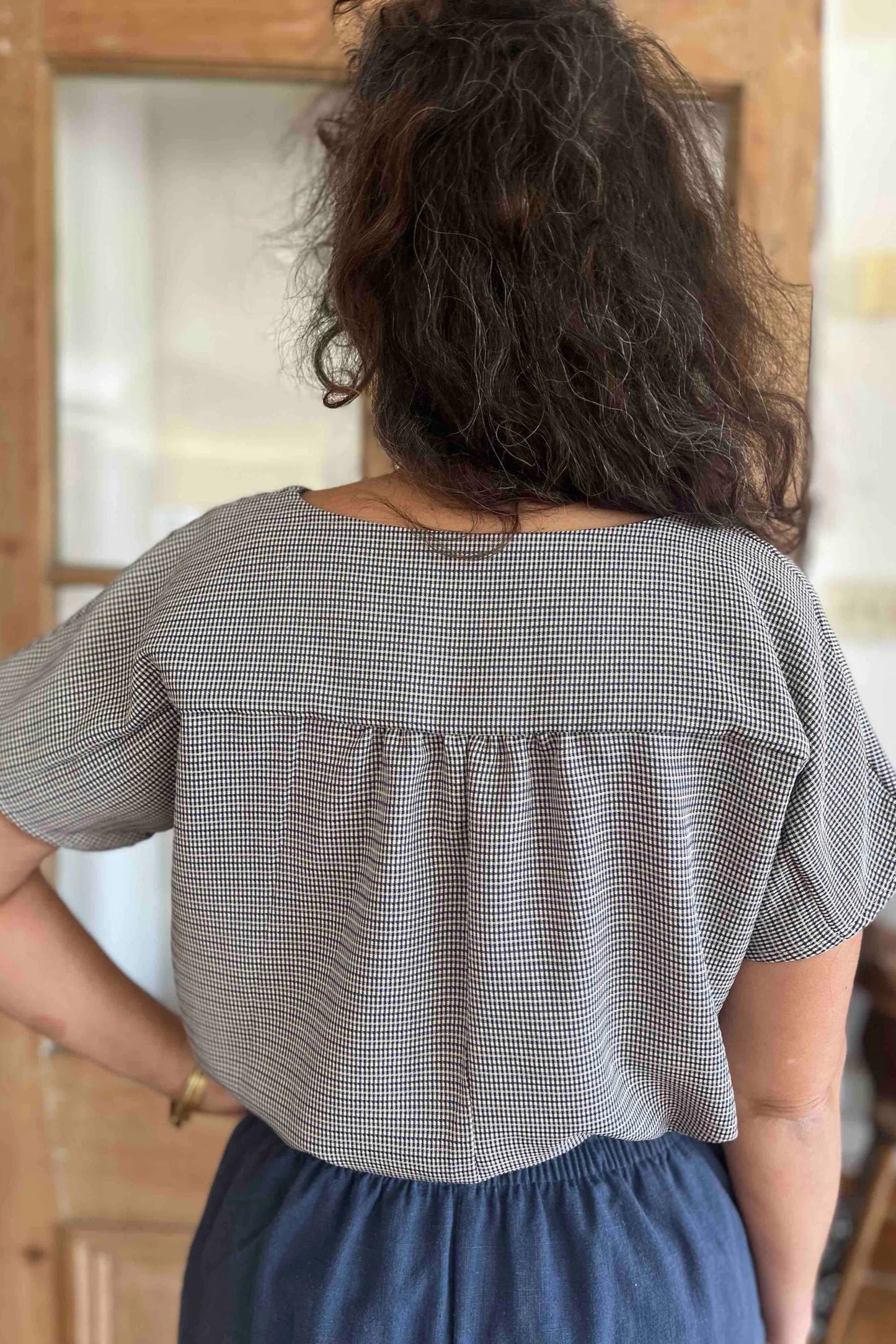 Back view of a woman wearing the Lucca Top in Blue Check print by Kazak, a short sleeved top with a notched V-neckline and a pleated yoke at the back. She is wearing it with the Sienna Pants in Lapis, and is standing in front of a wooden door.