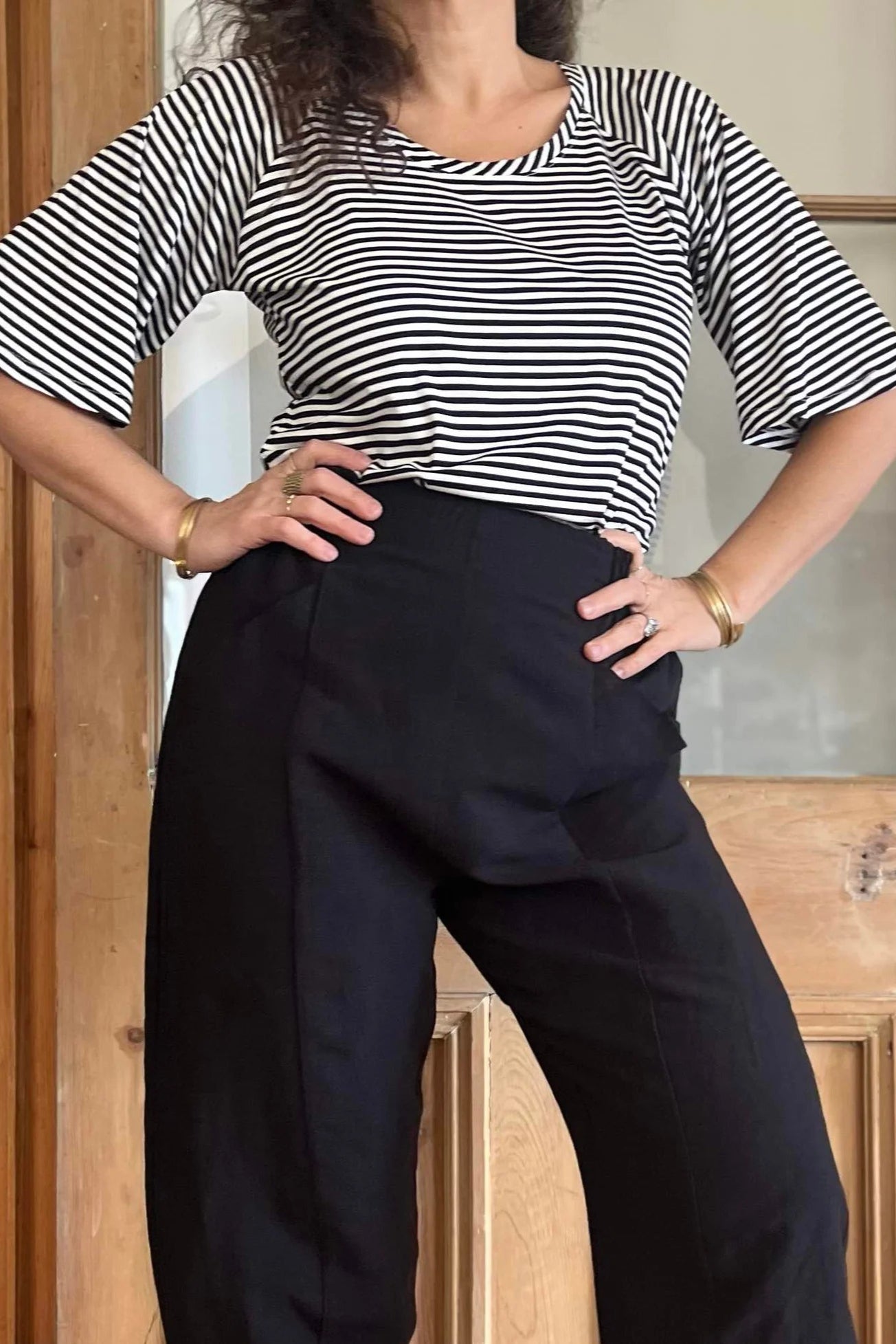 Neck down view of a woman wearing the Sienna Pants by Kazak in Black, featuring a flat front waistband with elastic at the sides and back, large angled pockets, and loose fitting legs that taper slightly at the ankle. She is wearing them with a striped top and is standing in front of a wooden door.
