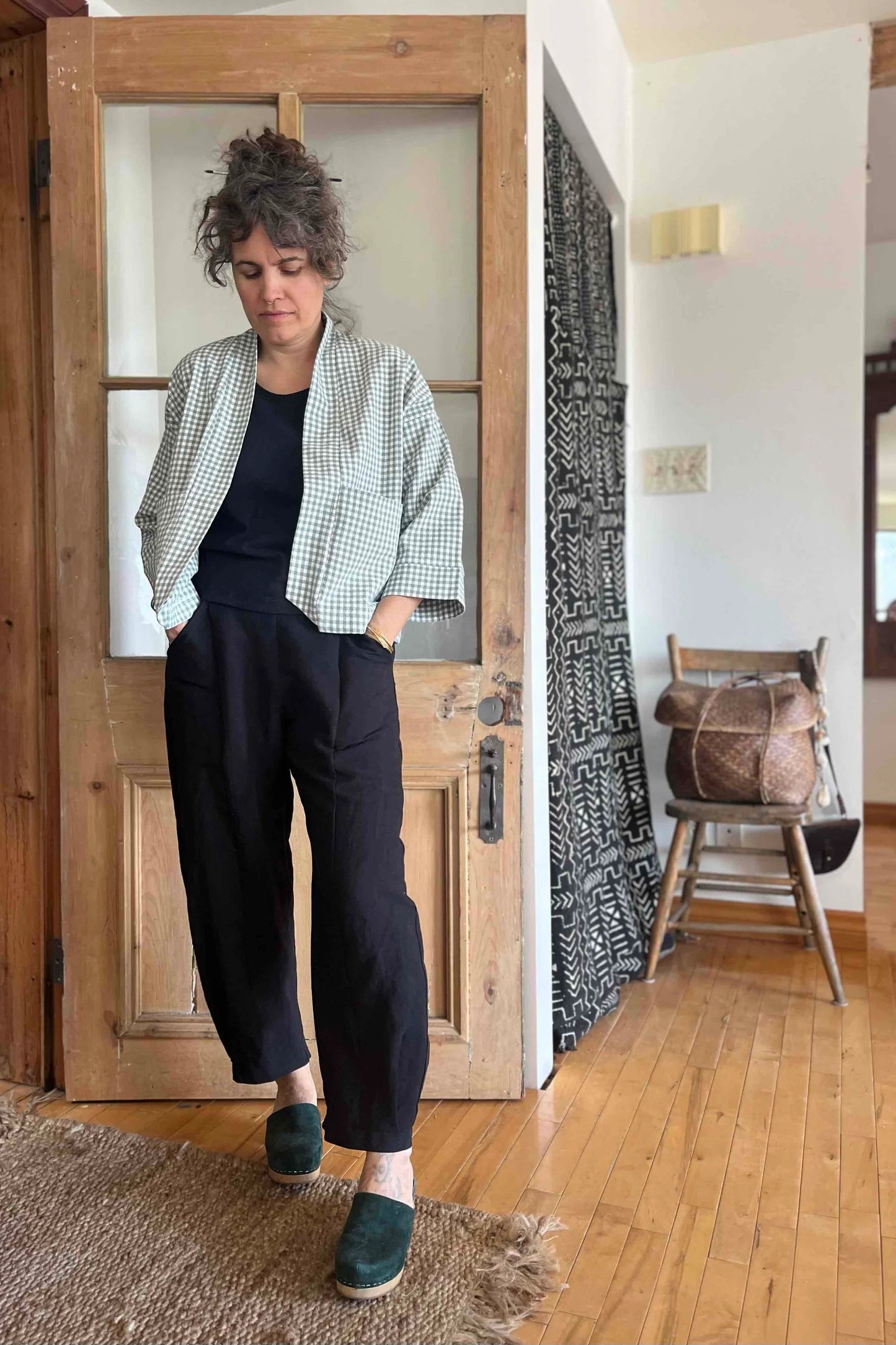 A woman wearing the Sienna Pants by Kazak in Black, featuring a flat front waistband with elastic at the sides and back, large angled pockets, and loose fitting legs that taper slightly at the ankle. She is wearing them with the a checked jacket and is standing in front of a wooden door.